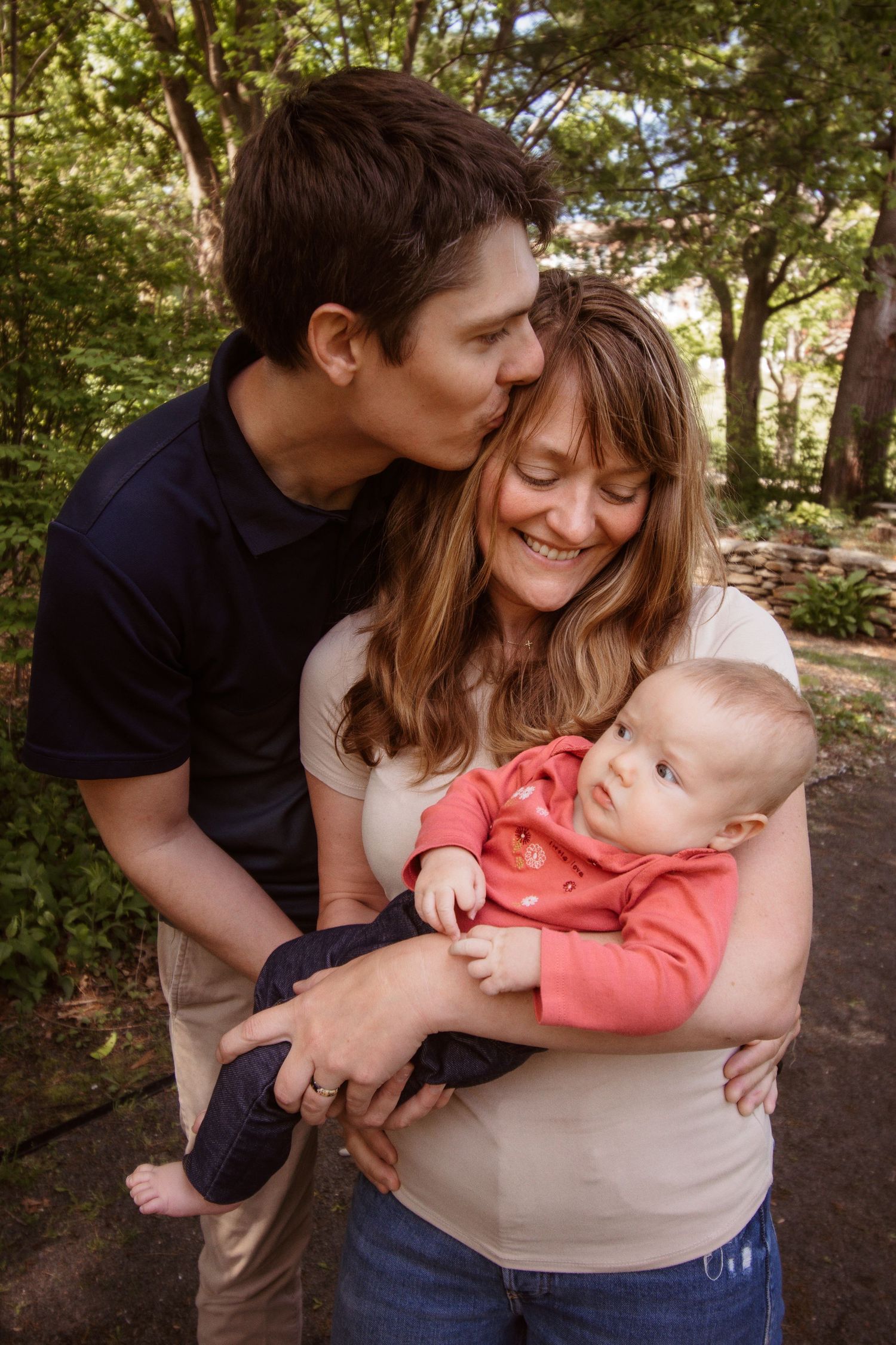 A couple outdoors holds and snuggles with their infant baby in a series of candid moments against a wooded background.