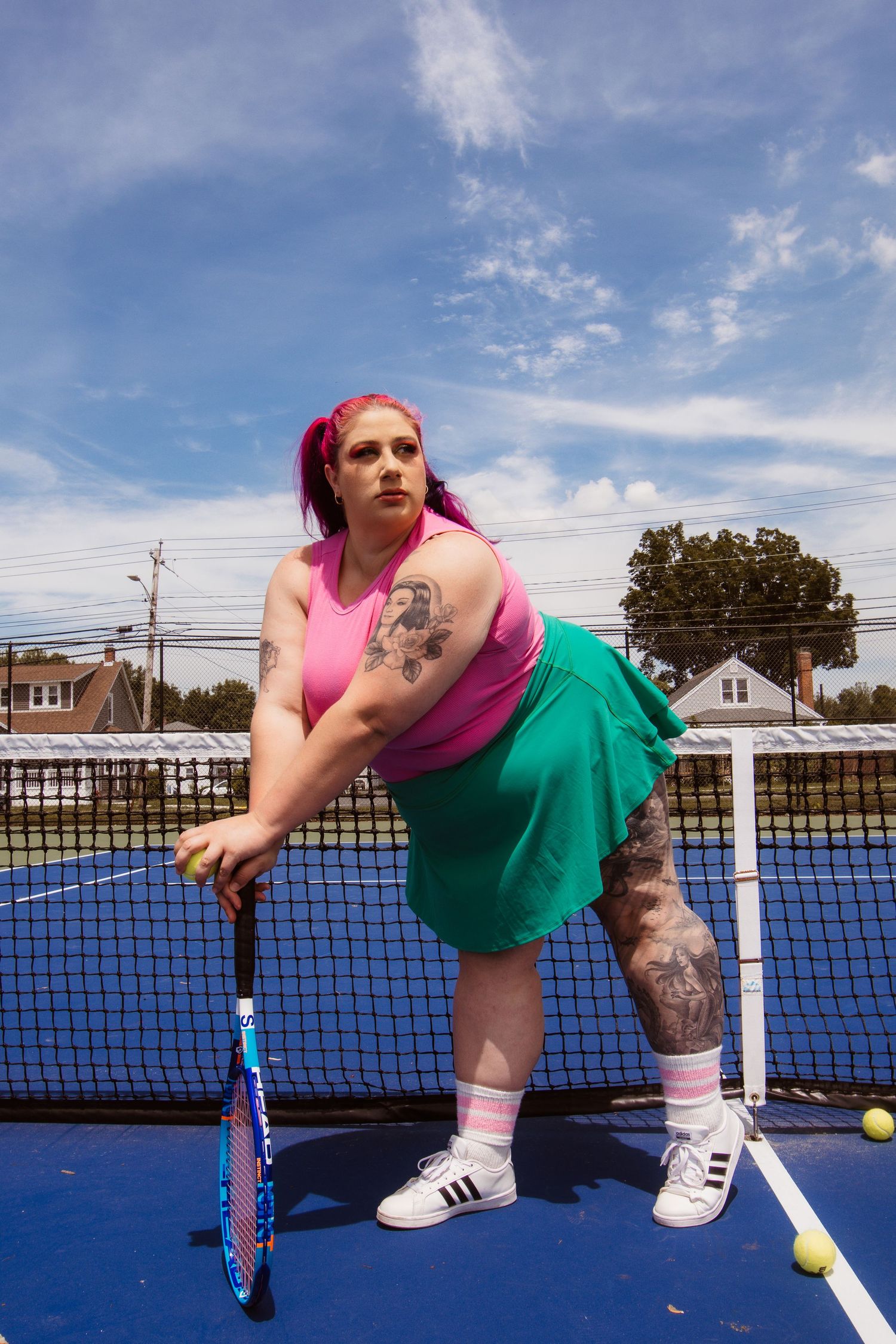 Person in pink top and green skirt stretches near tennis net on blue court under sunny sky.