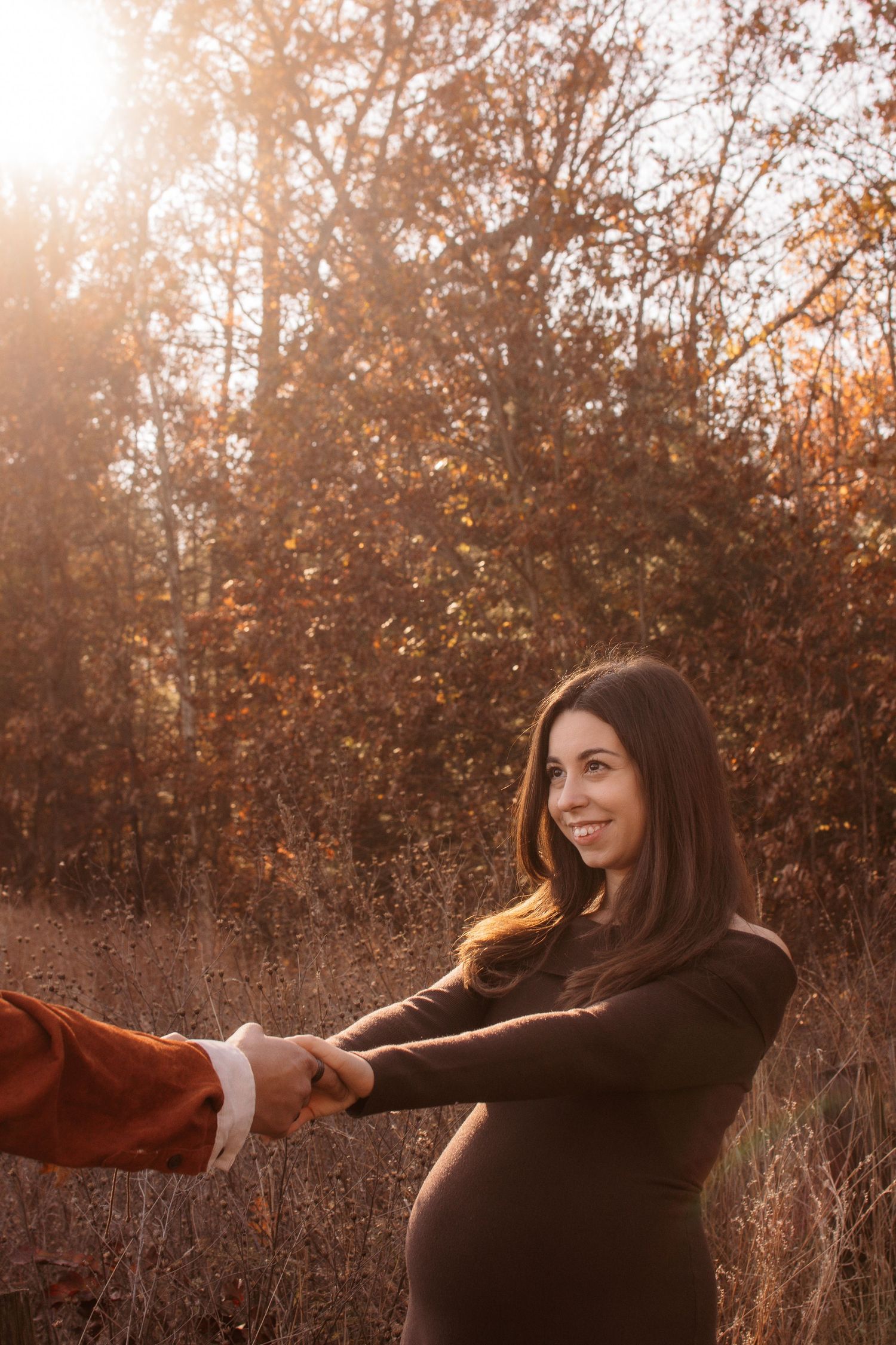 Woman in brown sweater twirls joyfully in autumn forest with golden sunlight filtering through trees.
