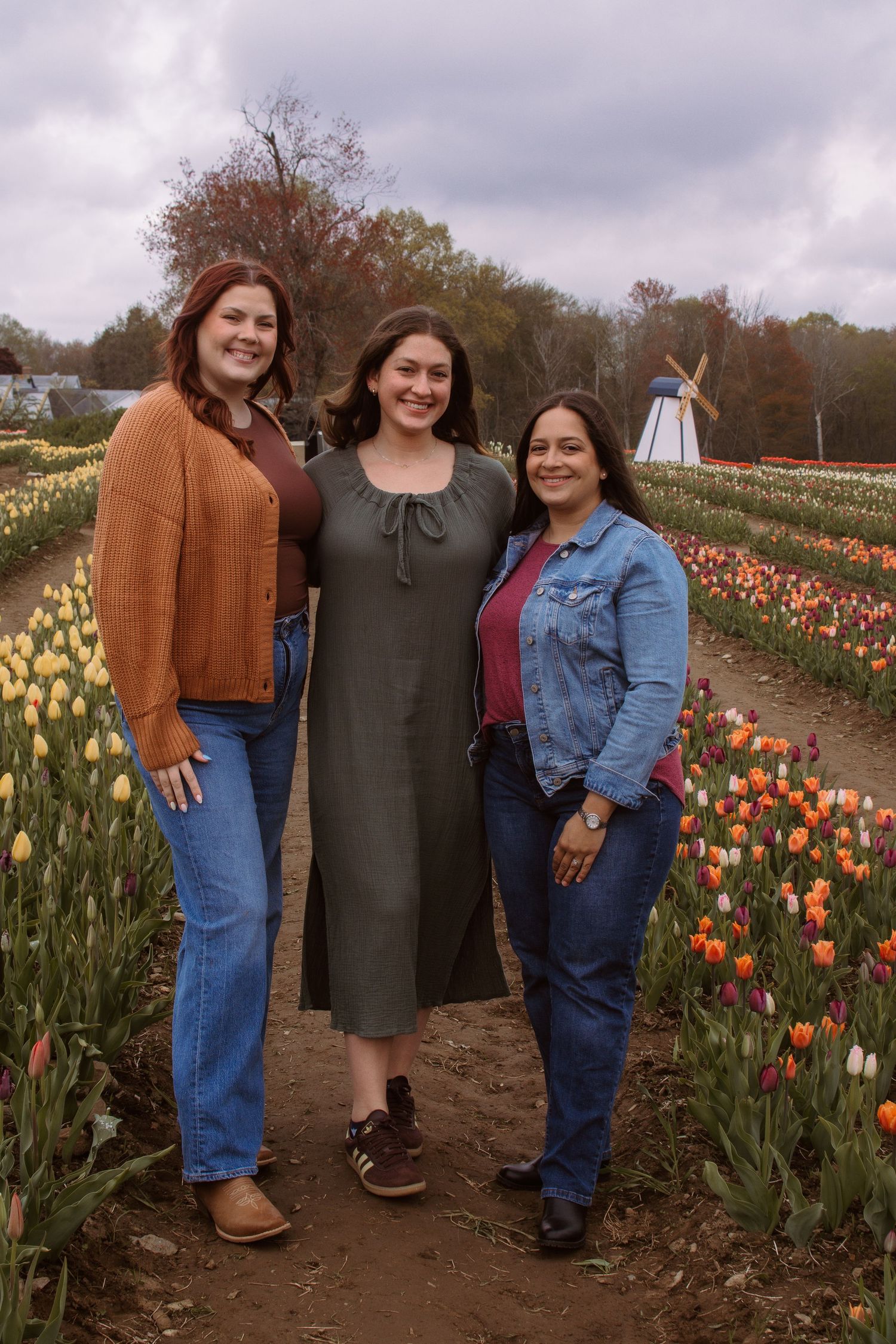 Three women smiling together in a colorful tulip field with a windmill in the background on a cloudy day.