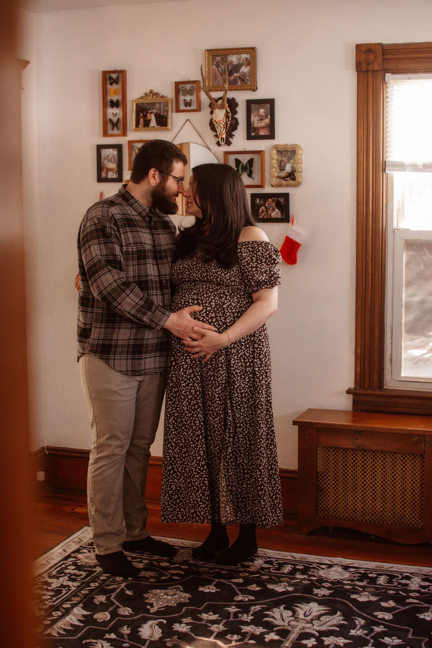 Couple sharing a tender moment, hands on pregnant belly, standing in a cozy home with a gallery wall and Christmas stocking.