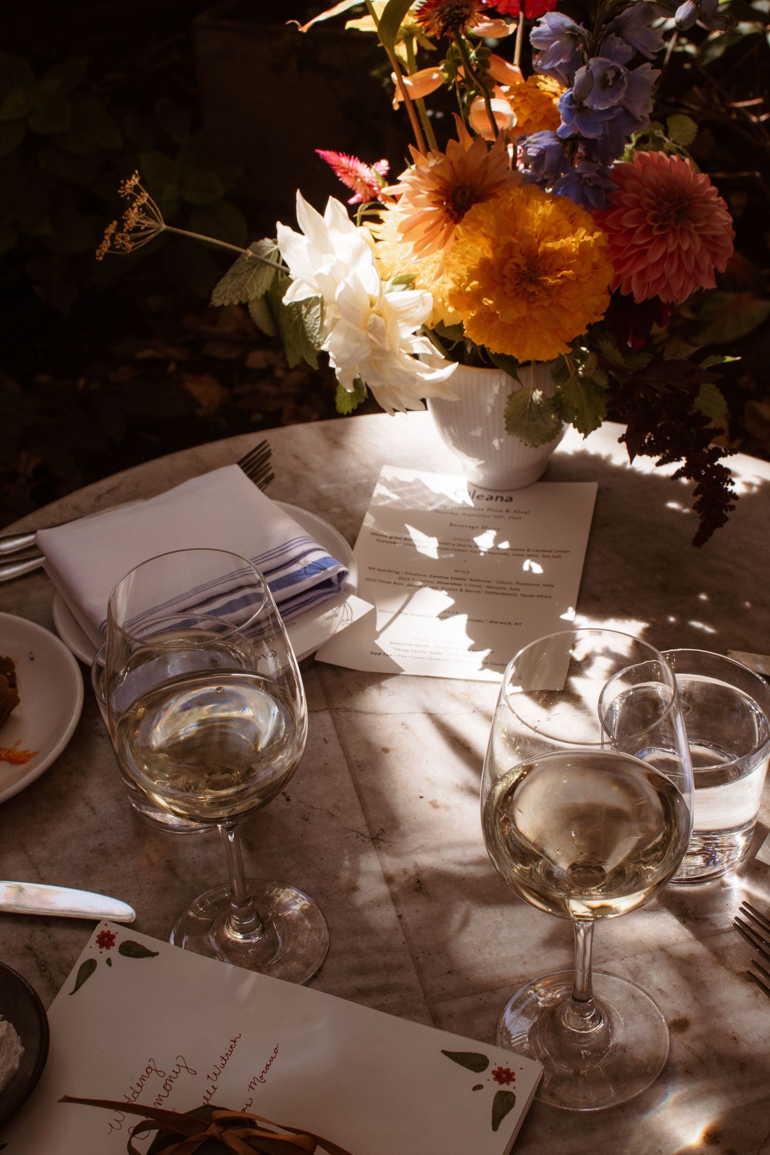 Wine glasses and floral centerpieces cast shadows on an elegant dining table setting in natural sunlight.