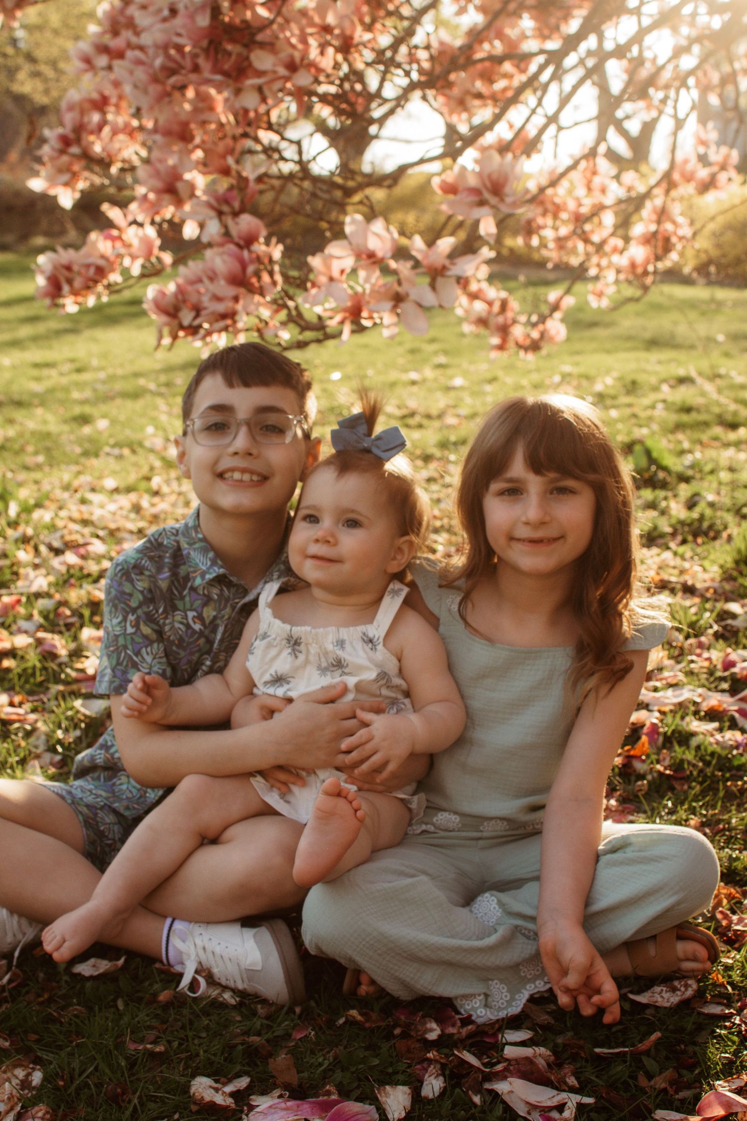 Three siblings sitting together on grass during golden hour with cherry blossoms blooming overhead in spring.