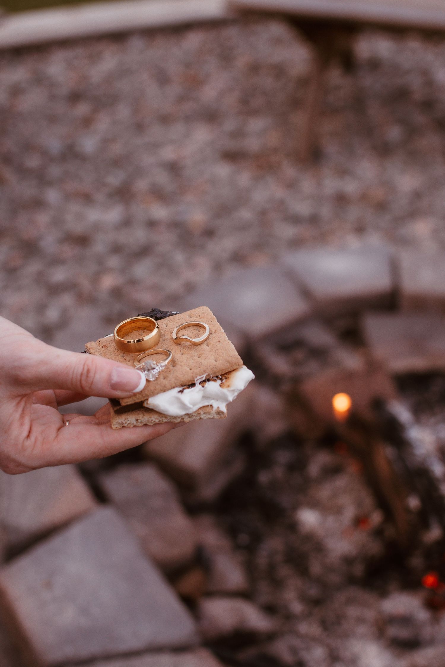 Hand holding a s'more with two gold wedding rings on top near a glowing campfire.