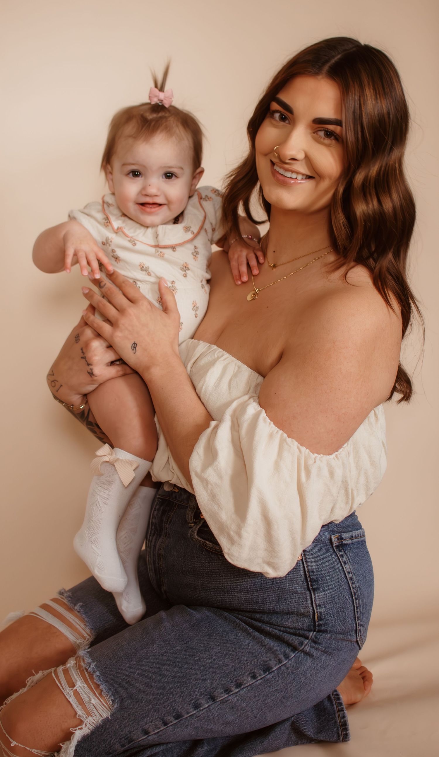 Against a beige backdrop, someone in a white peasant top and blue jeans poses with a small infant in white clothing.