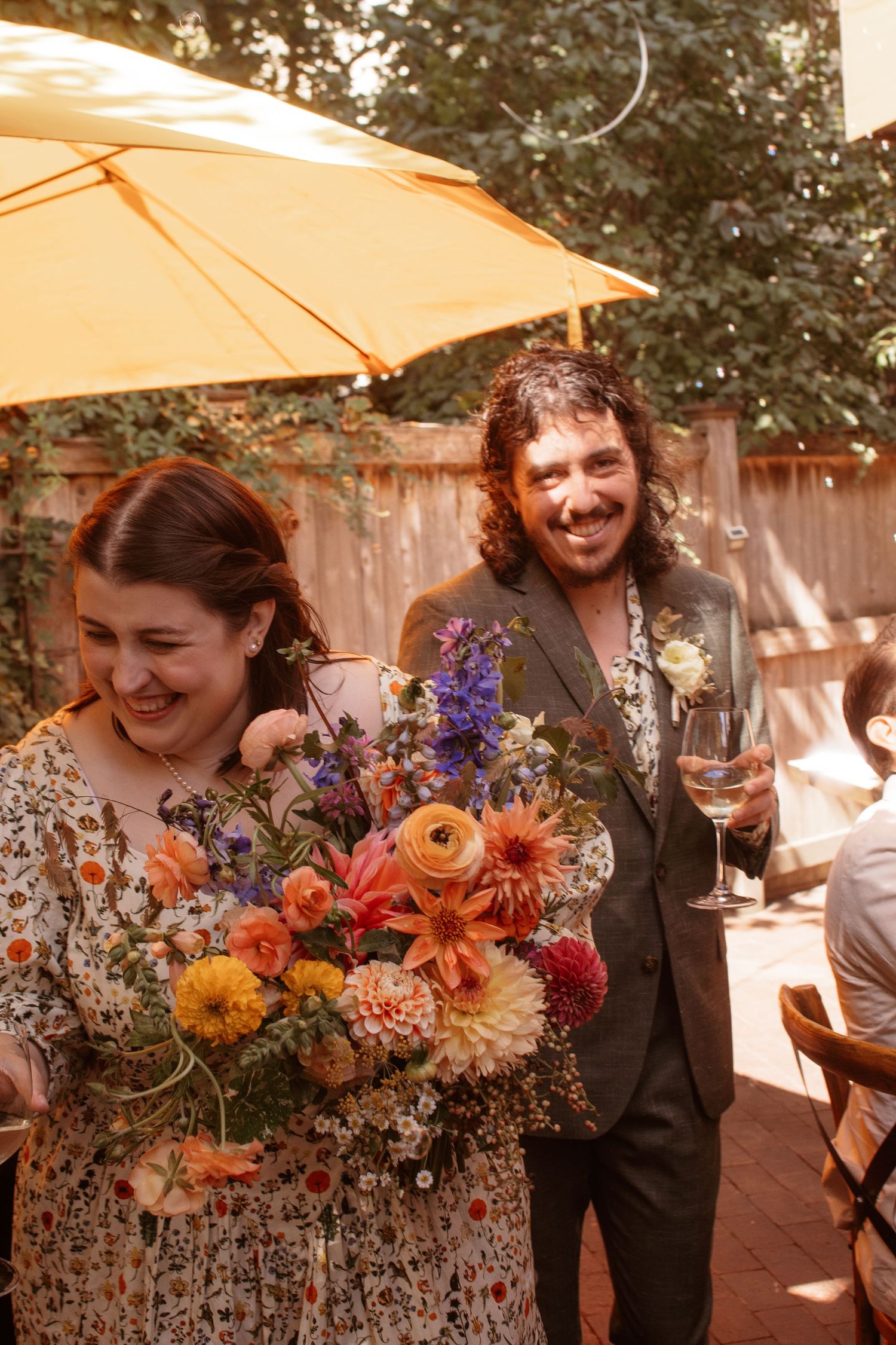 A couple shares joyful moments at an outdoor celebration with vibrant orange and purple floral bouquets.