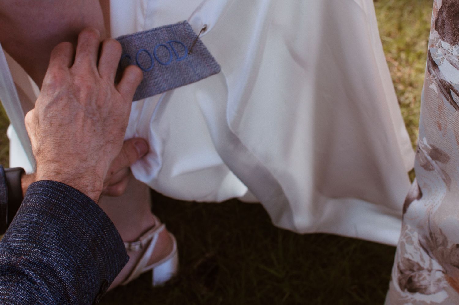 Close-up of hands pinning a blue tag onto a white wedding dress outdoors.