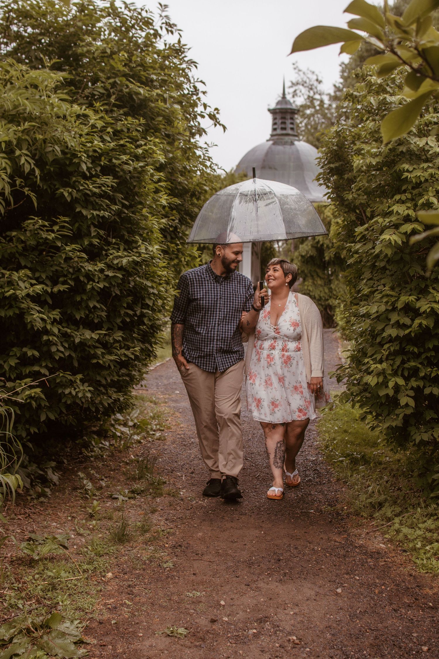 Couple walking under clear umbrella on garden path, smiling at each other, lush greenery and dome building behind.