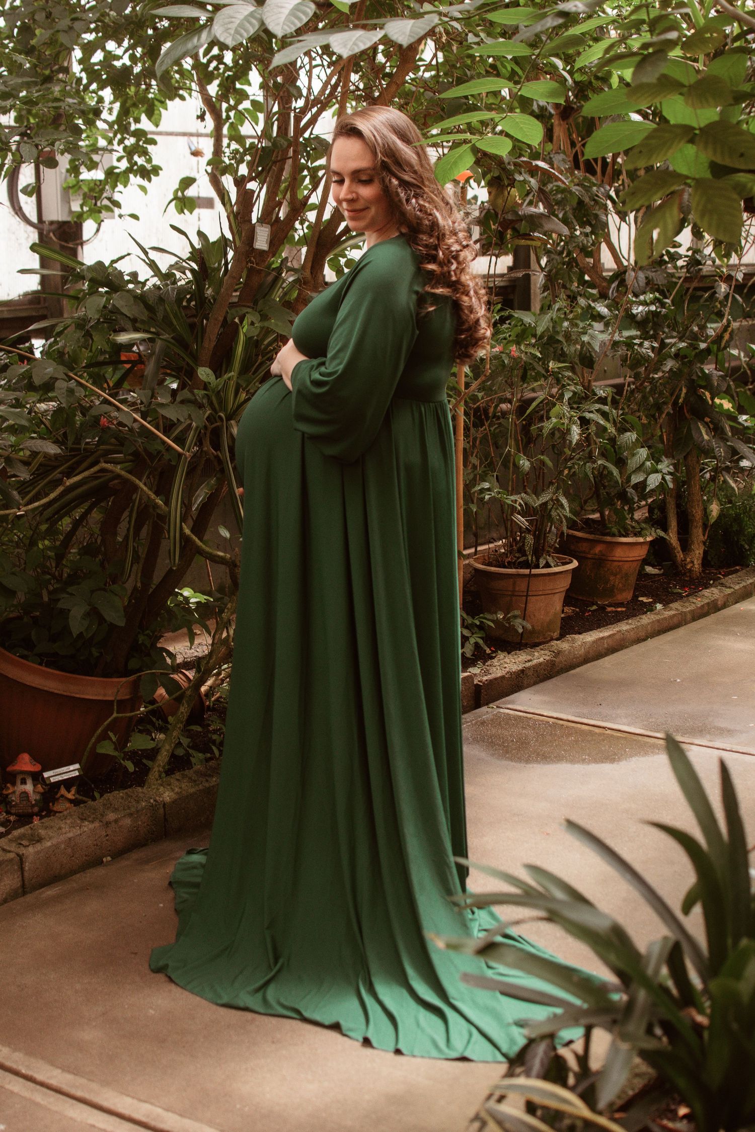 A figure in a flowing emerald green gown poses gracefully among potted plants and greenery in a greenhouse setting.