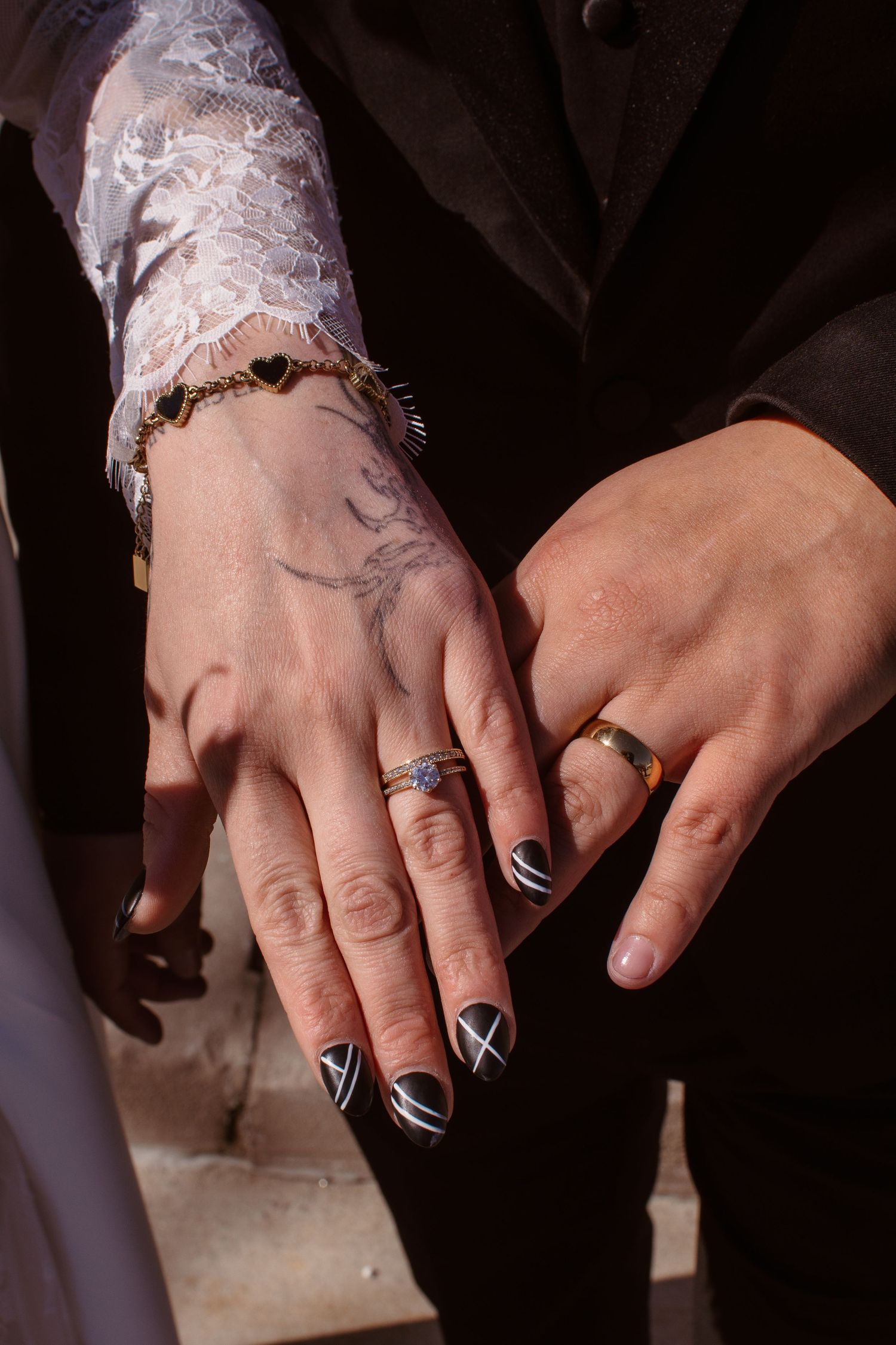Two newlyweds display wedding rings, one hand with black geometric nails, tattoo, and bracelet, the other with a gold band.