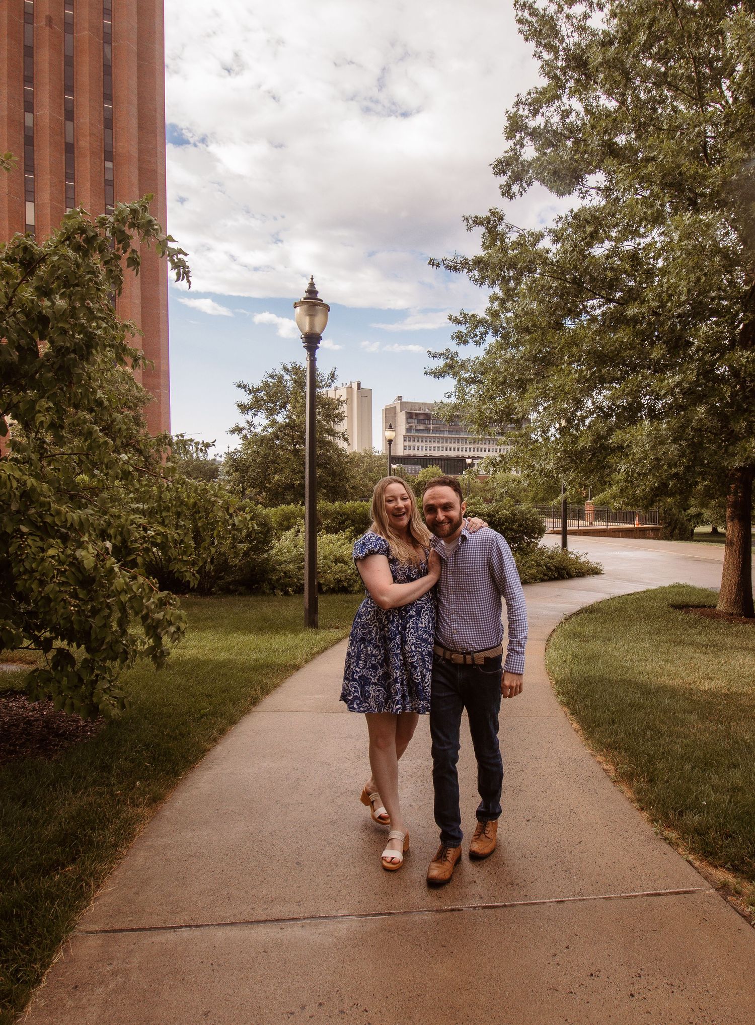 A smiling couple poses on a tree-lined campus pathway near a vintage lamppost on a cloudy summer day.