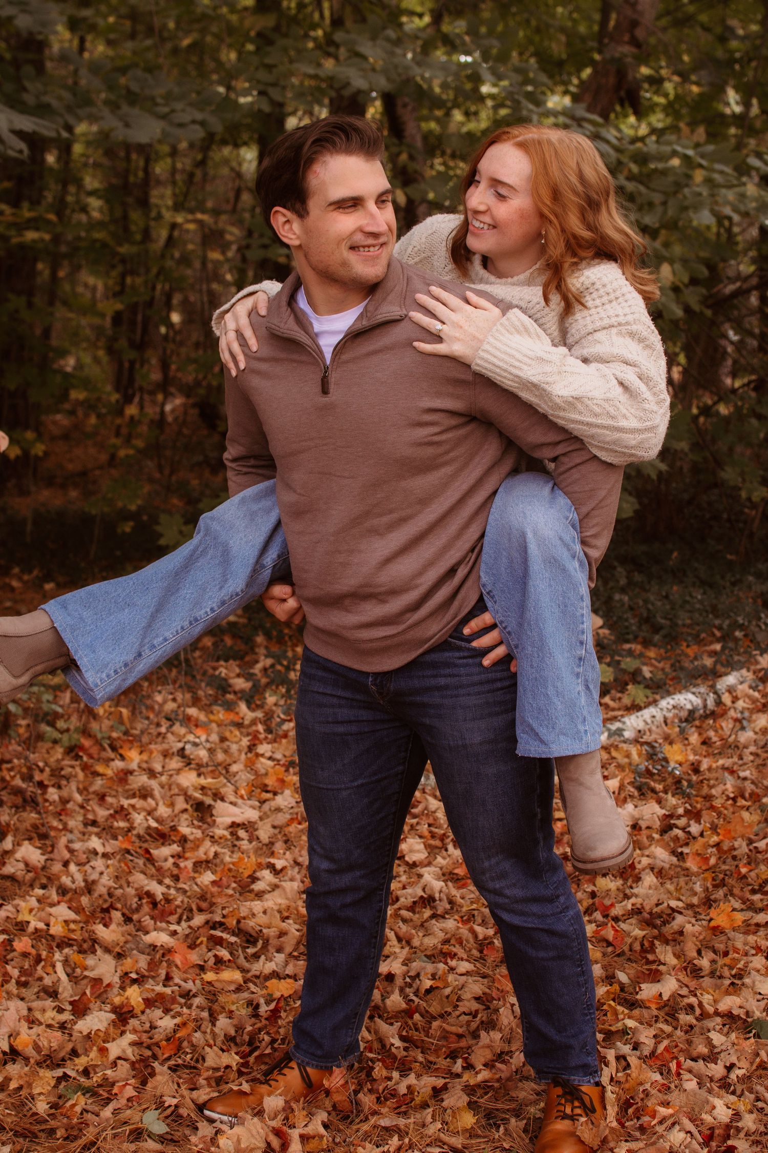 Playful autumn engagement photos with couple in brown and cream sweaters among fallen leaves.