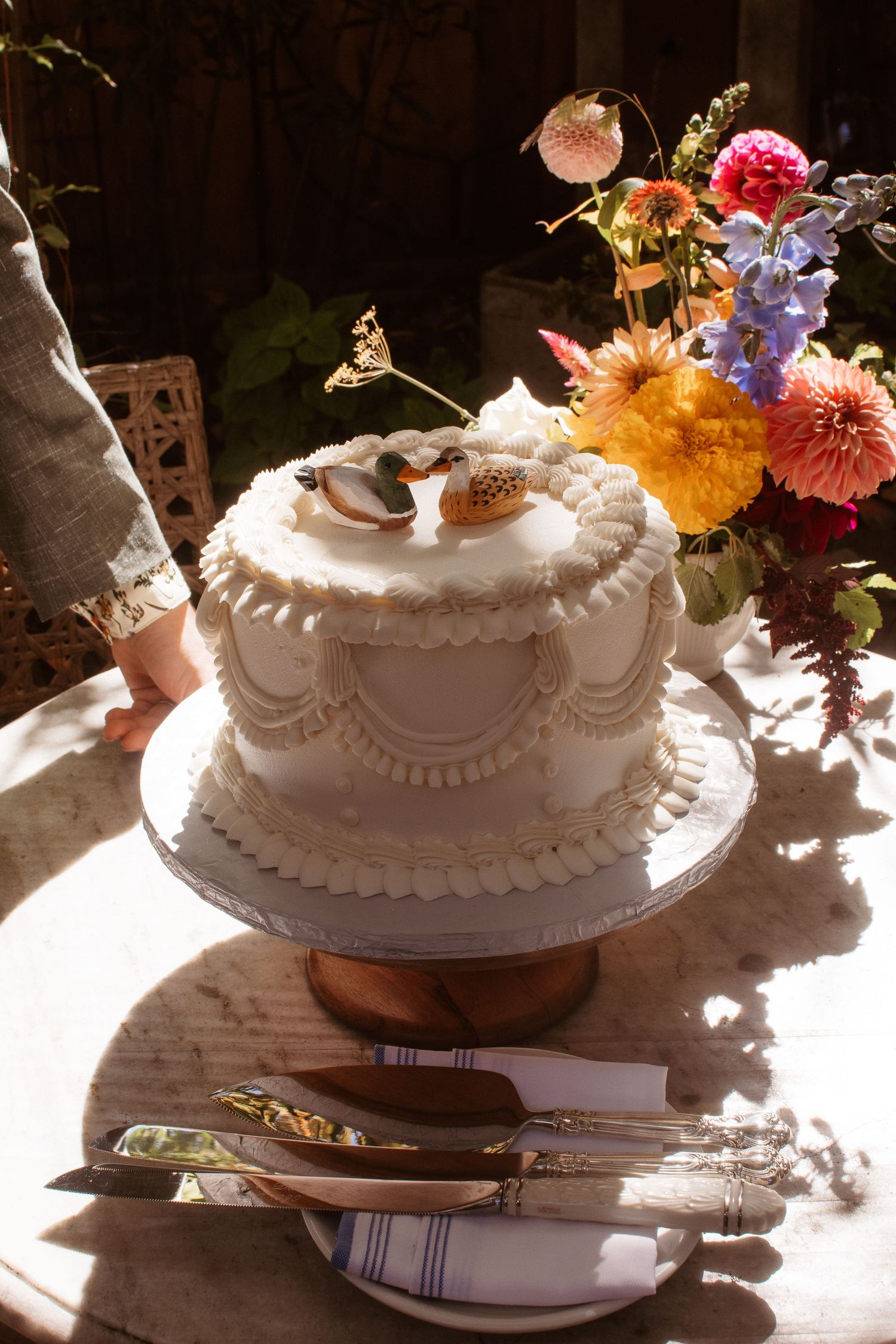 A white wedding cake with elegant piping sits on a cake stand next to colorful floral arrangements in sunlit setting.