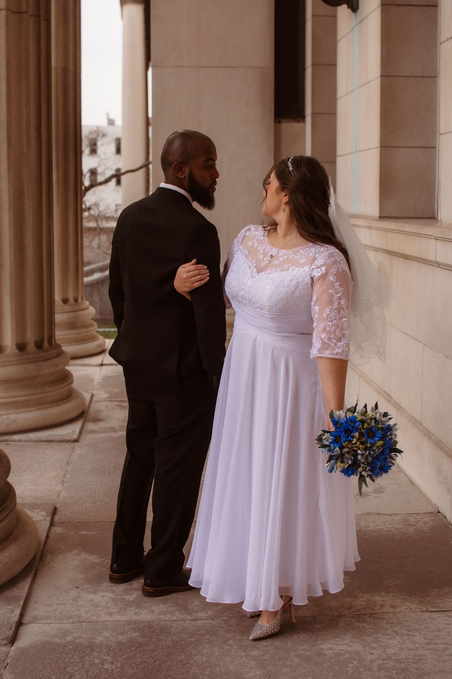 Romantic wedding couple shares intimate moments in a classic columned corridor with a blue floral bouquet.