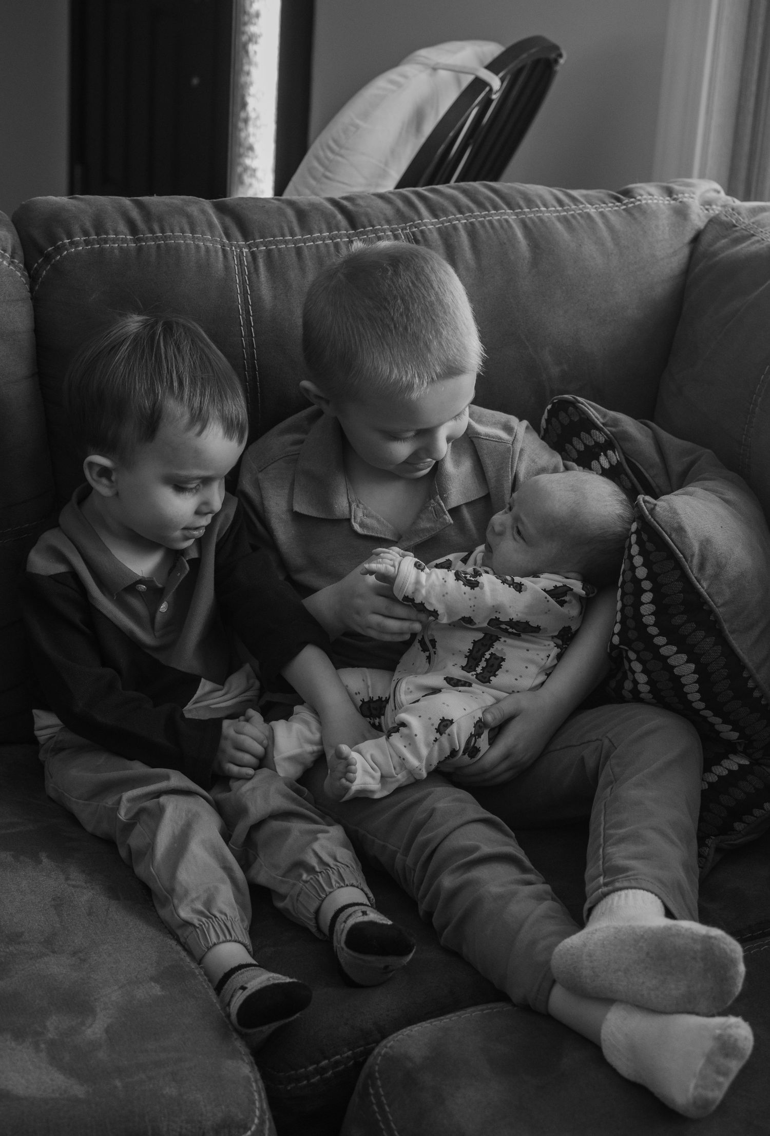 Black and white photo of three young siblings sitting together on a couch, older boys holding a newborn baby.