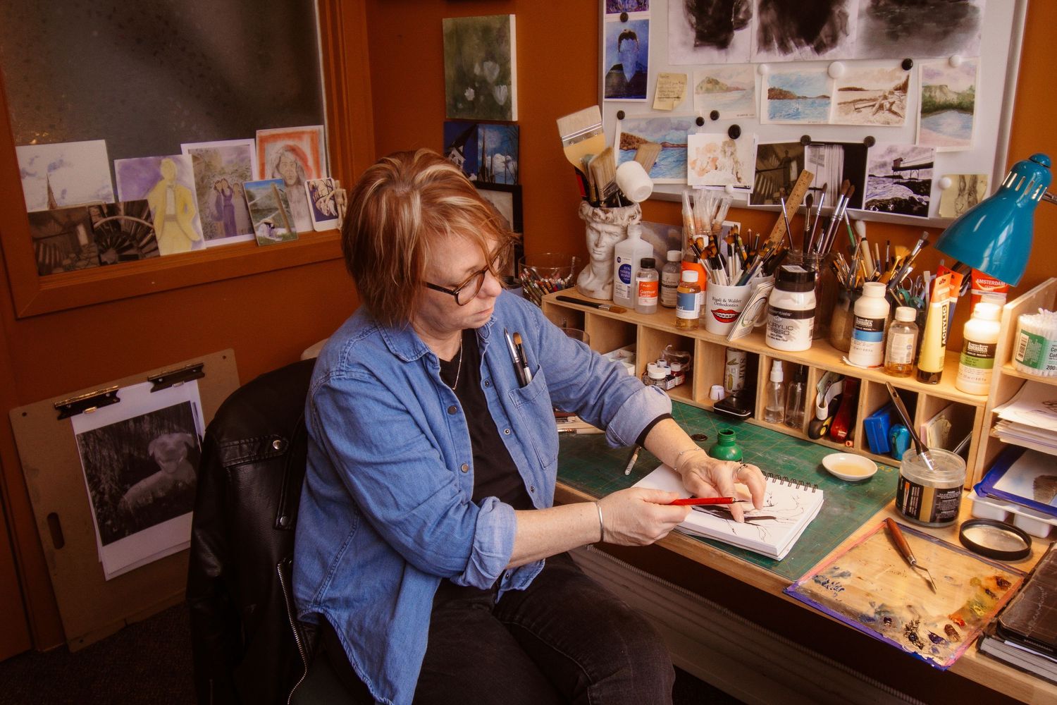 Artist working at a cluttered studio desk, painting with brushes and art supplies on shelves in a cozy creative workspace.