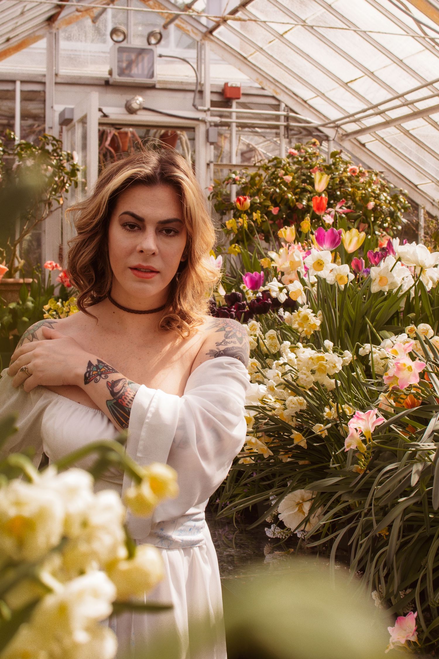 Woman in white dress with tattoos poses among colorful tulips and daffodils in a sunlit greenhouse.