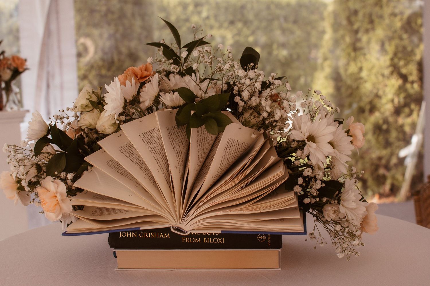 Elegant wedding centerpiece with an open book fanned out, surrounded by white and peach flowers on stacked John Grisham novels.