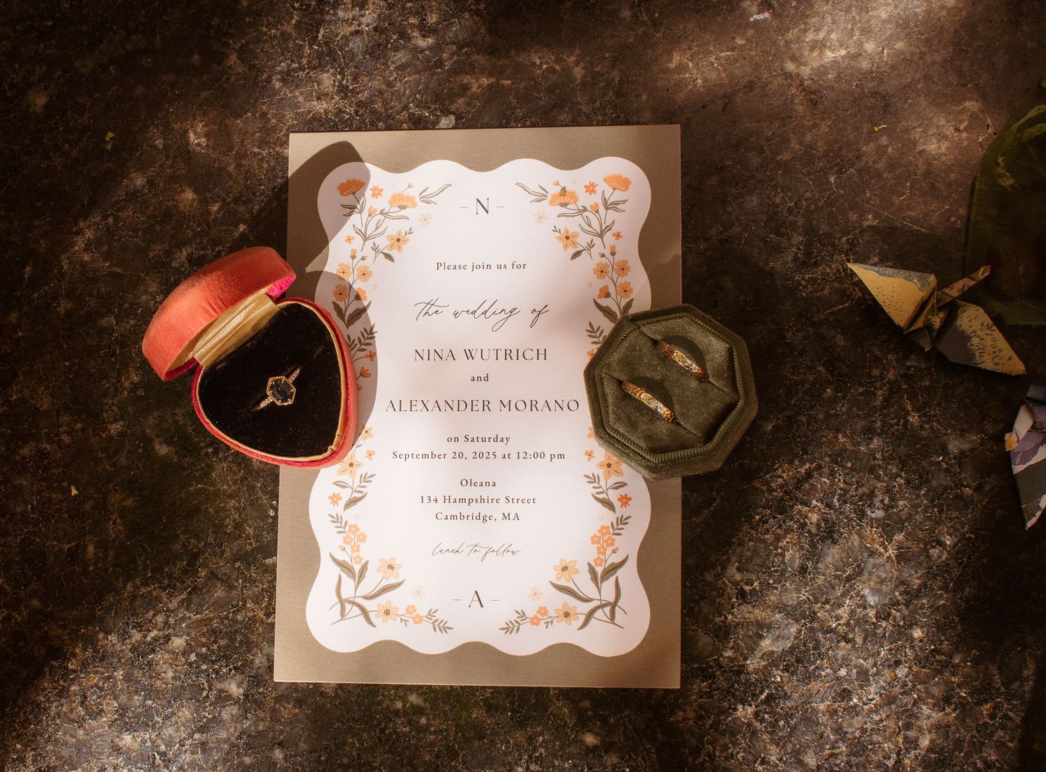 Wedding invitation with elegant gold floral design displayed alongside ring boxes on dark textured surface.