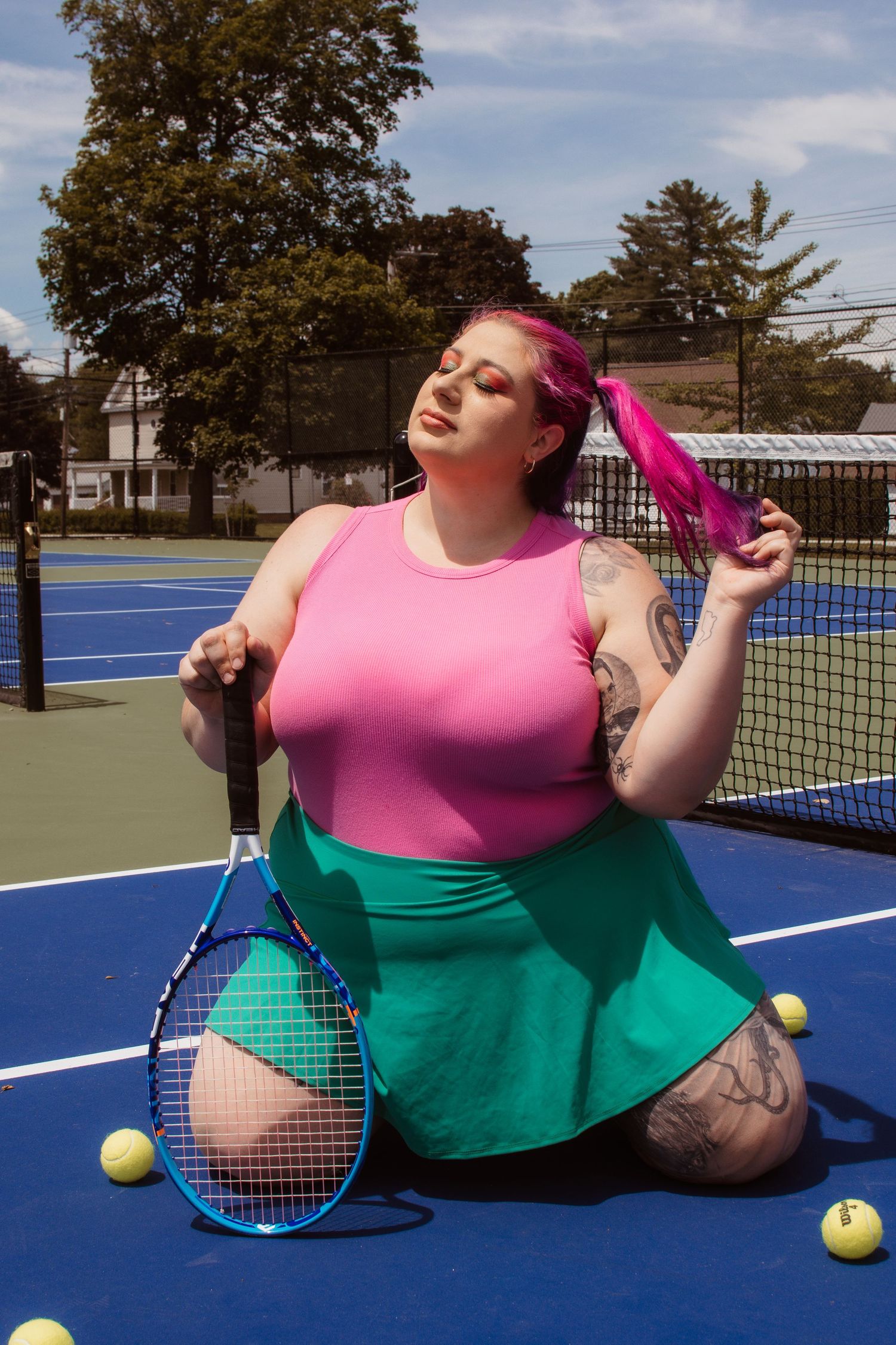 Tennis player in pink tank top and green skirt poses on blue court with racquet and tennis balls.