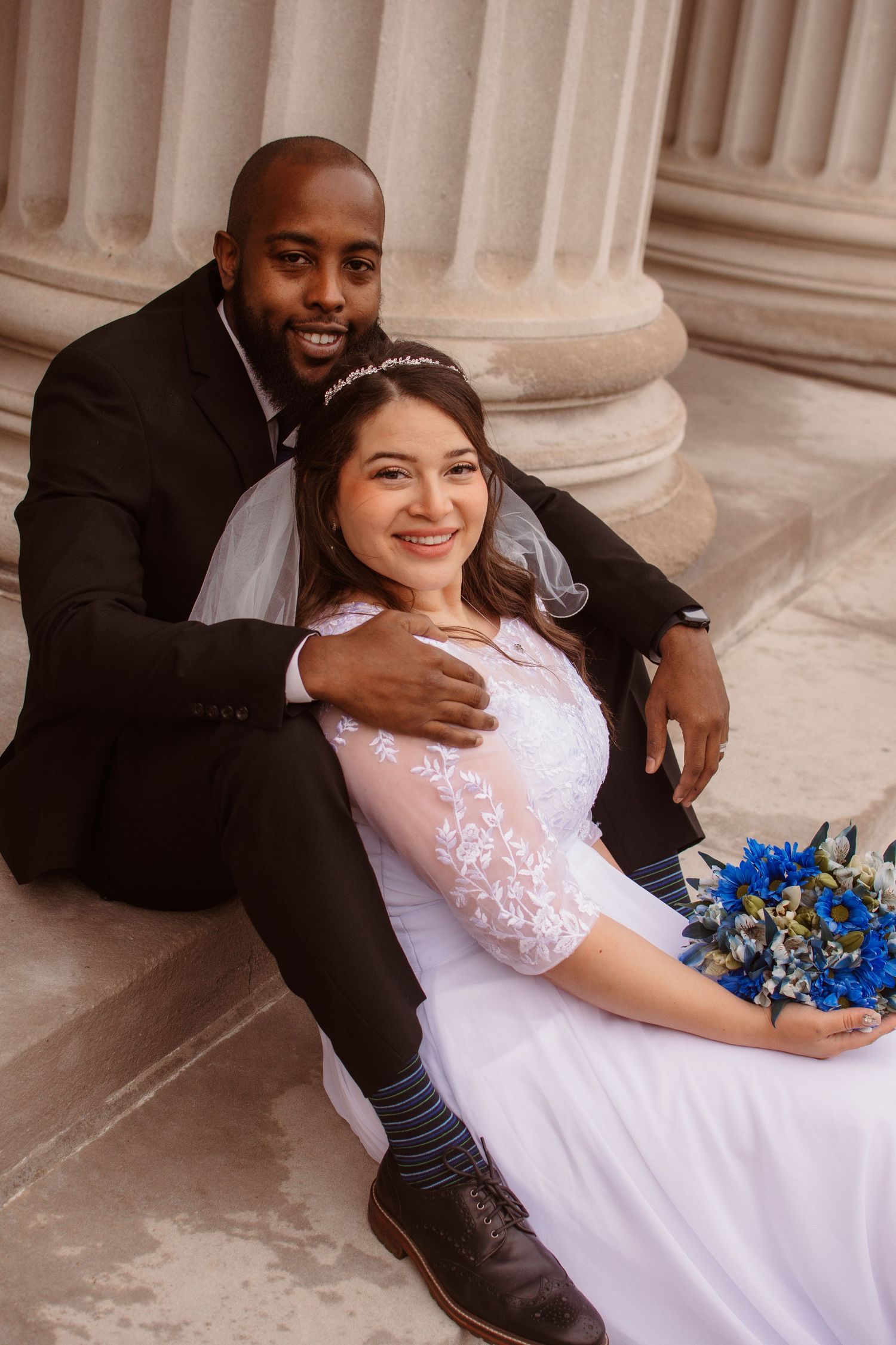 A bride in a white lace dress holding blue flowers poses with her groom on stone steps near classic columns.