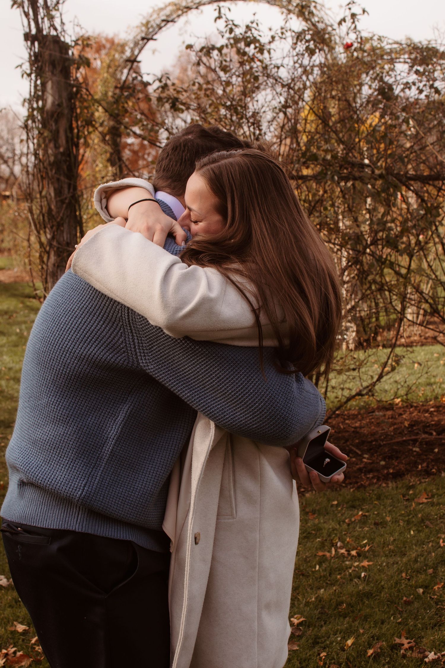 A romantic couple embraces outdoors during autumn with fallen leaves and trees in the background.