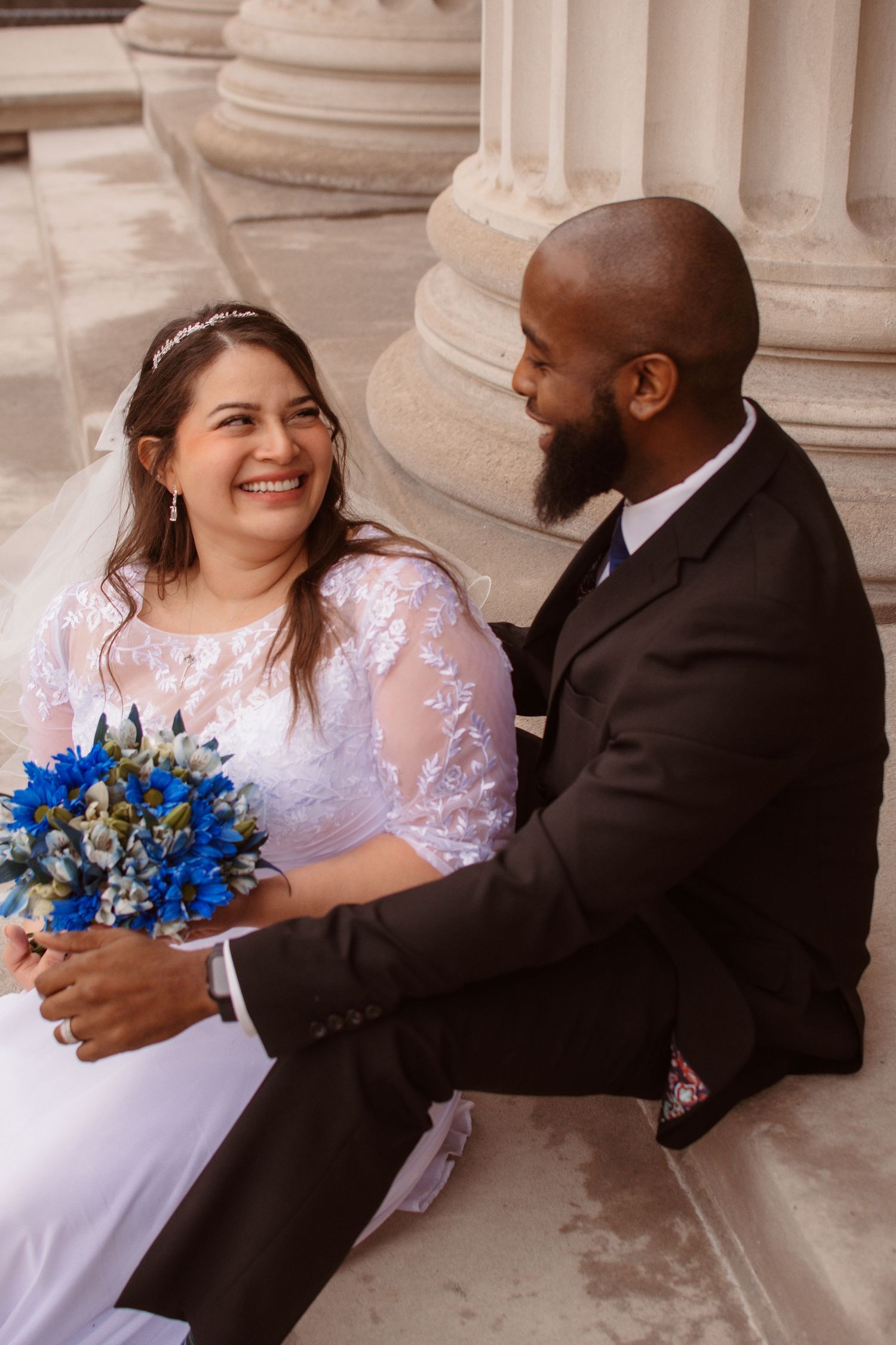 A wedding couple sits on marble steps sharing intimate moments with blue floral bouquet and white lace dress.