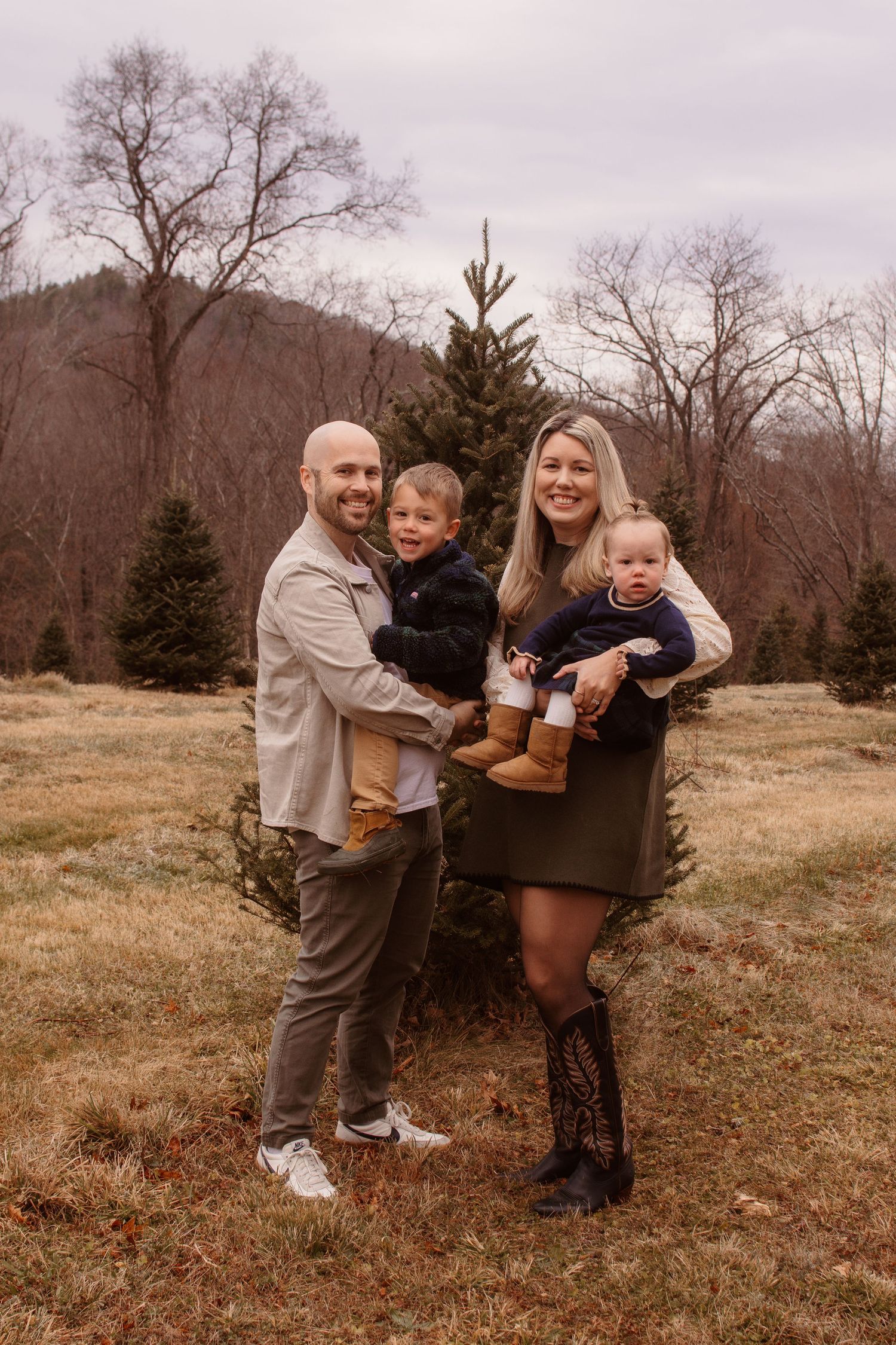 A family poses together in a rustic autumn setting with bare trees and evergreens in the mountainous background.