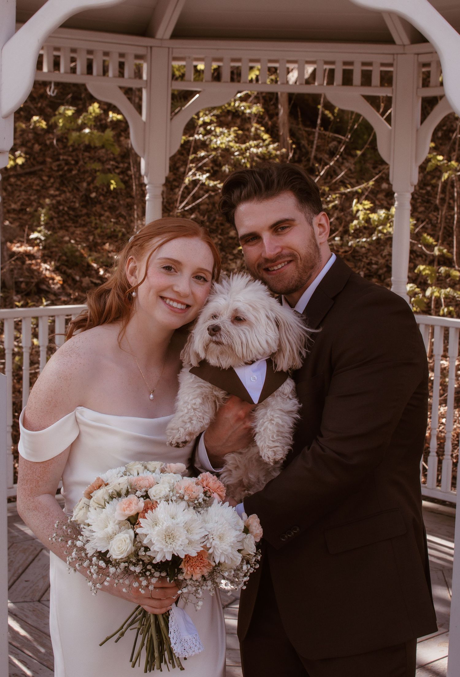 Bride and groom holding a white fluffy dog in a tuxedo, posing under a gazebo with a bridal bouquet.