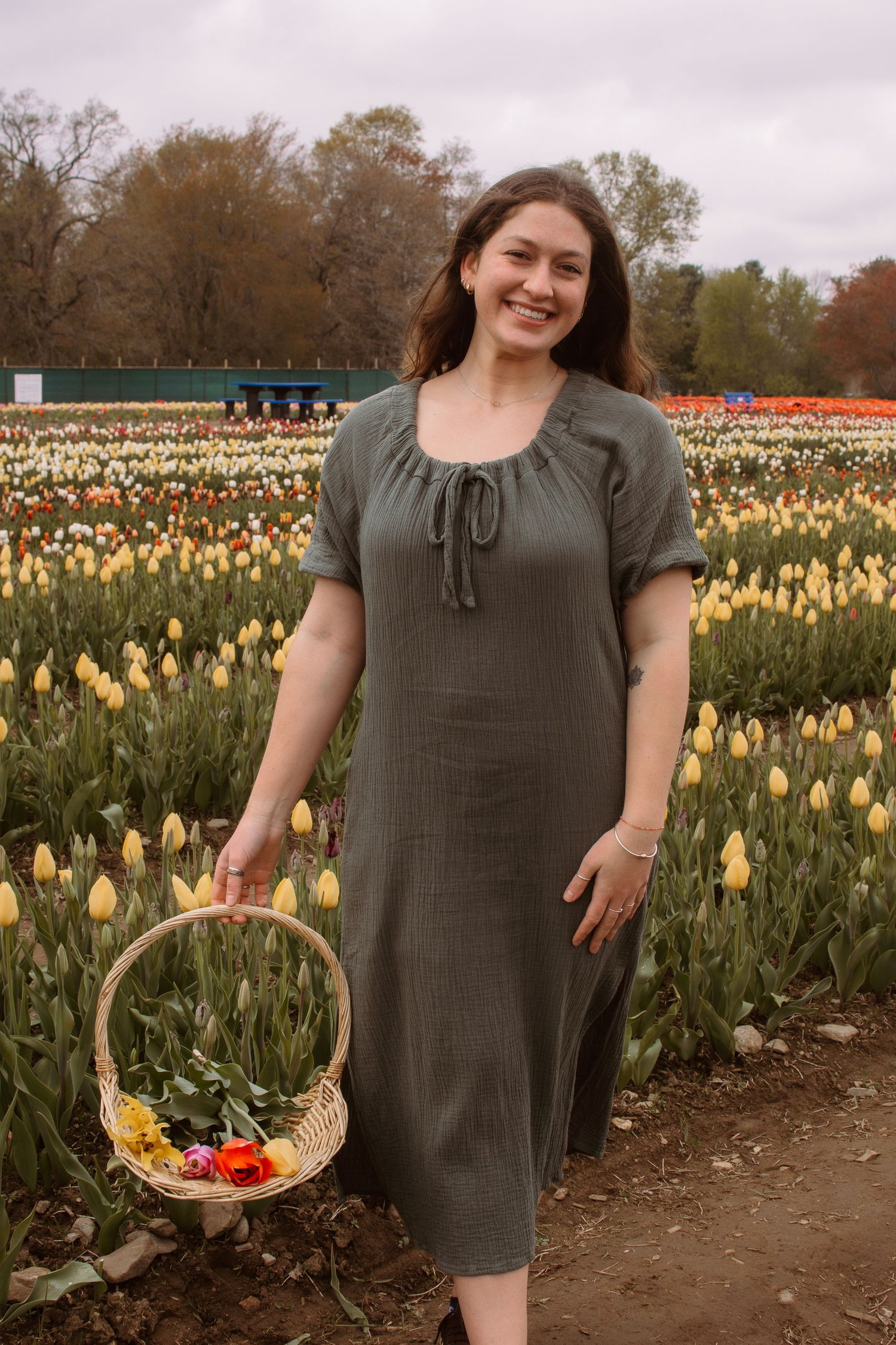 Smiling woman in olive green dress holding a wicker basket of flowers in a colorful tulip field.