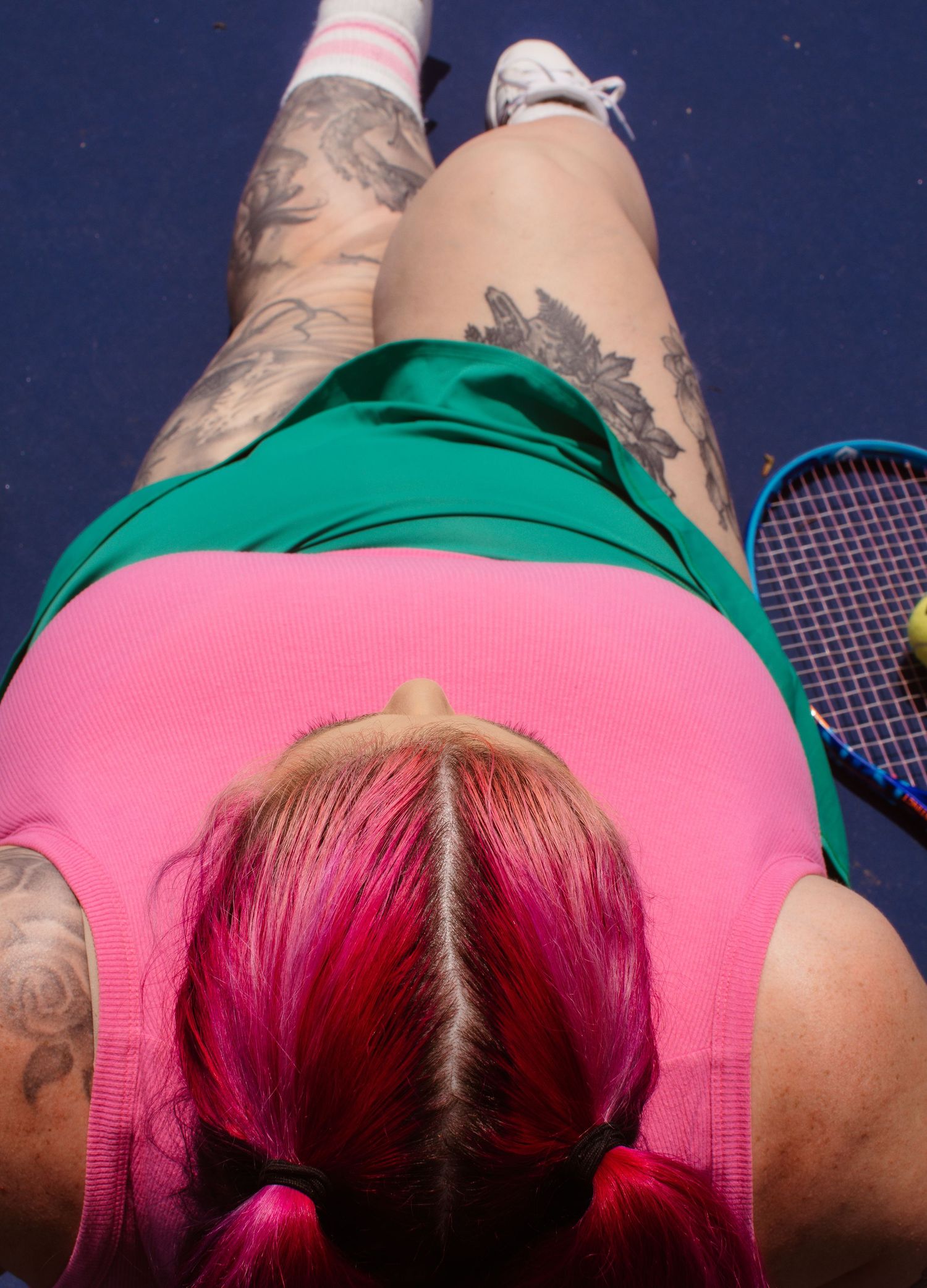 An overhead view of a tennis player wearing a green top and pink shorts with visible tattoos on their legs and arms.