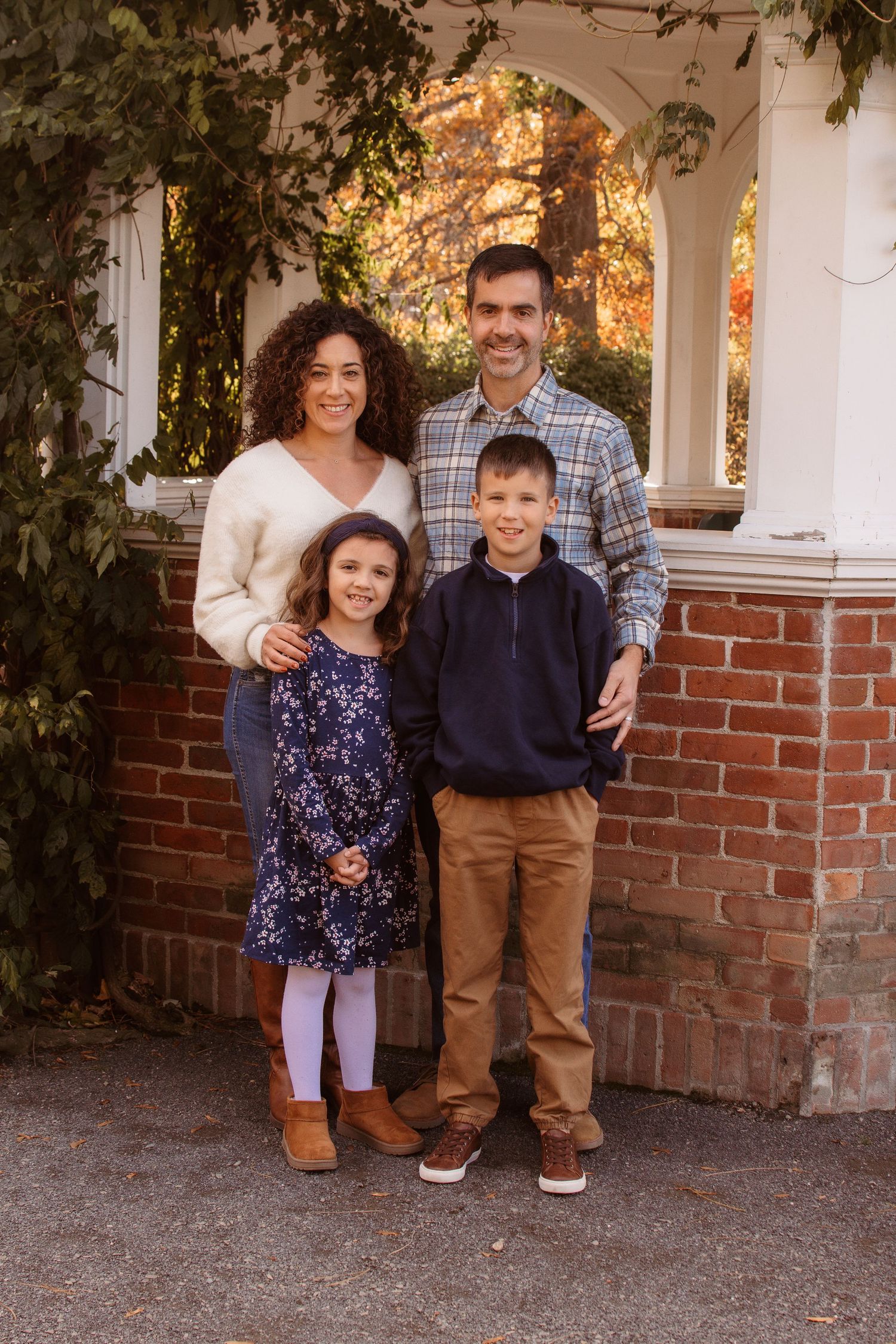 A family of four poses together on an autumn day in front of a brick porch with white columns and climbing vines.