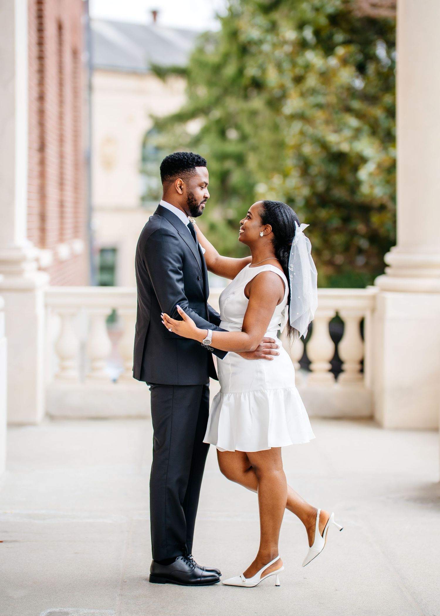 A couple in formal wear embraces on an elegant balcony with classic architectural details.