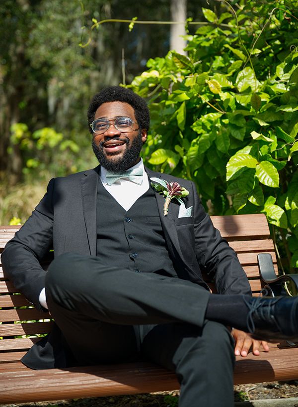 Groom in classic black tuxedo relaxing on wooden bench in garden setting.