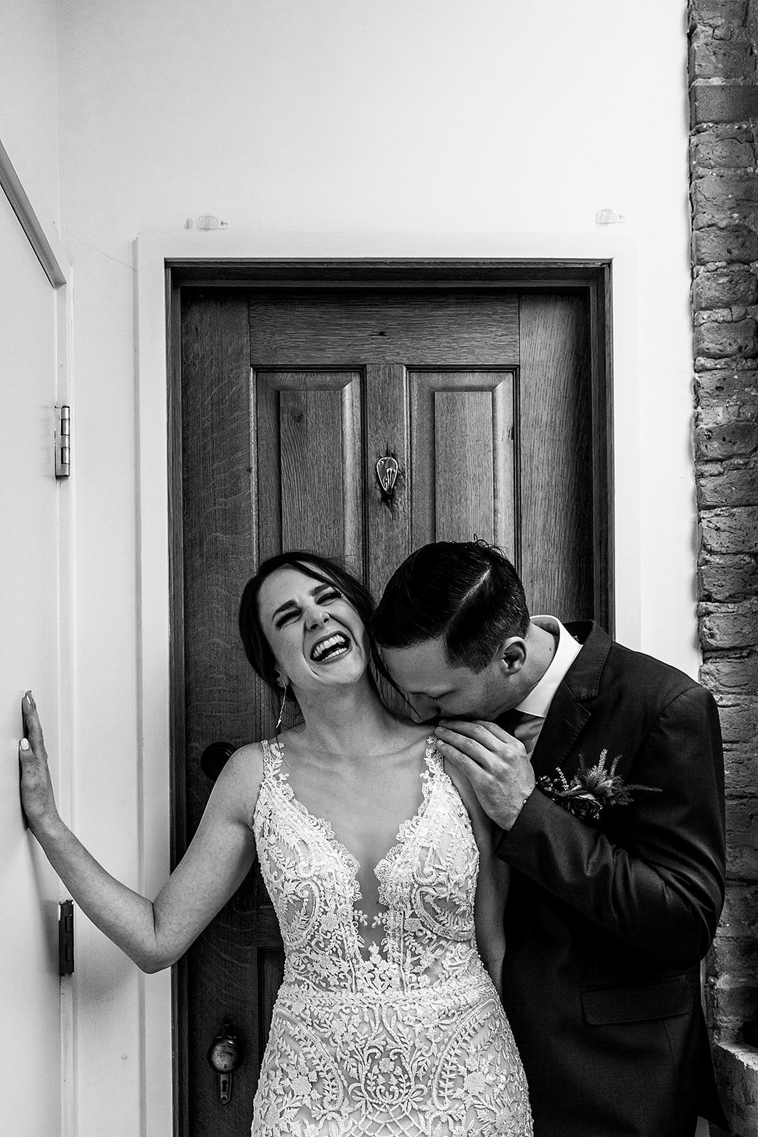 Black and white candid wedding photo captured against wooden door showing joyful intimate moment.