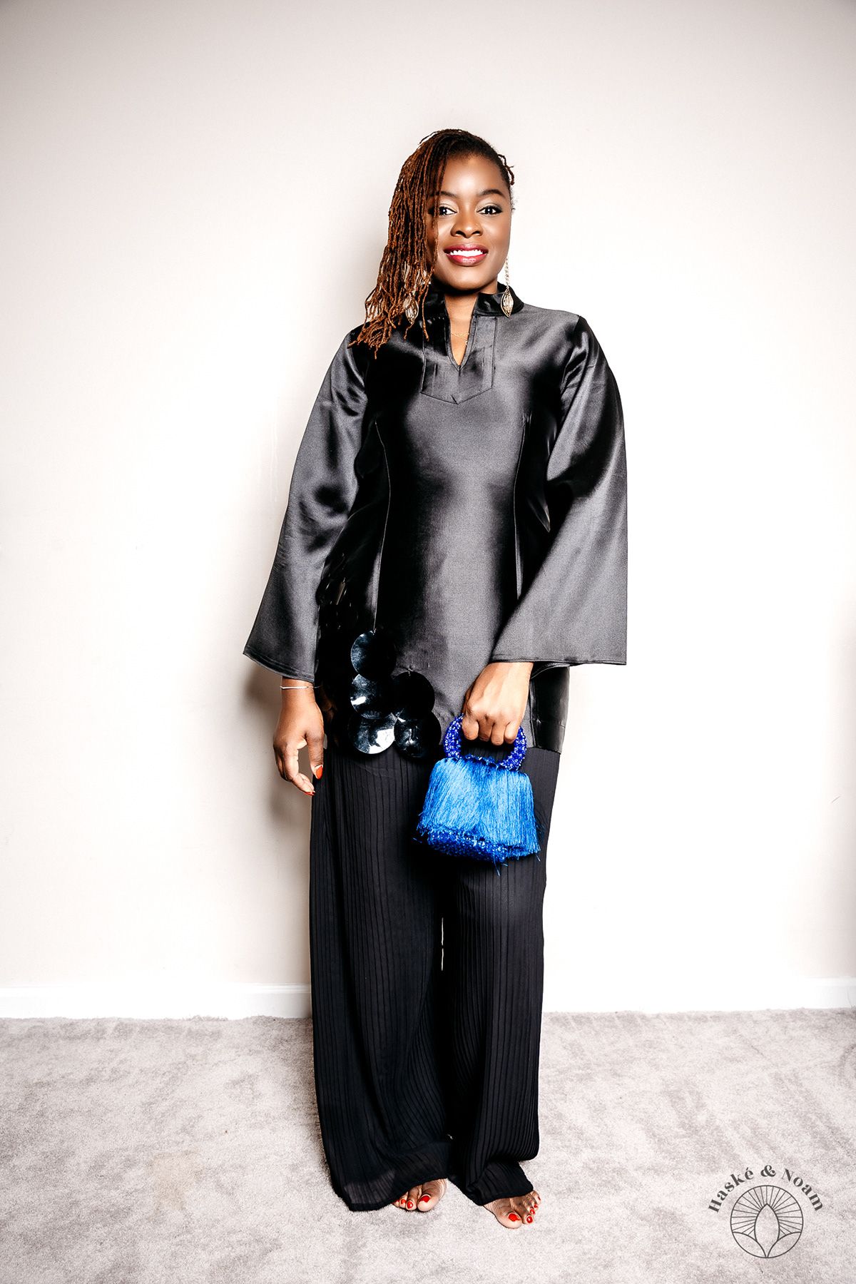A person in a black silk top and wide-leg pants holding a small blue handbag against a white background.