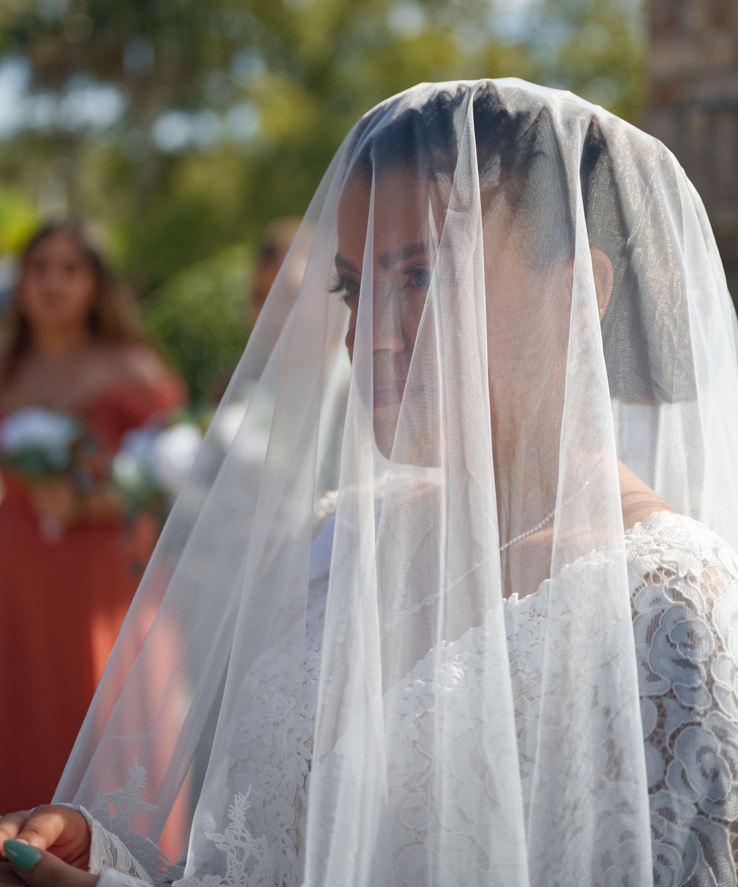 A bride wears a sheer white veil over her lace wedding dress while a bridesmaid in coral stands in the background.