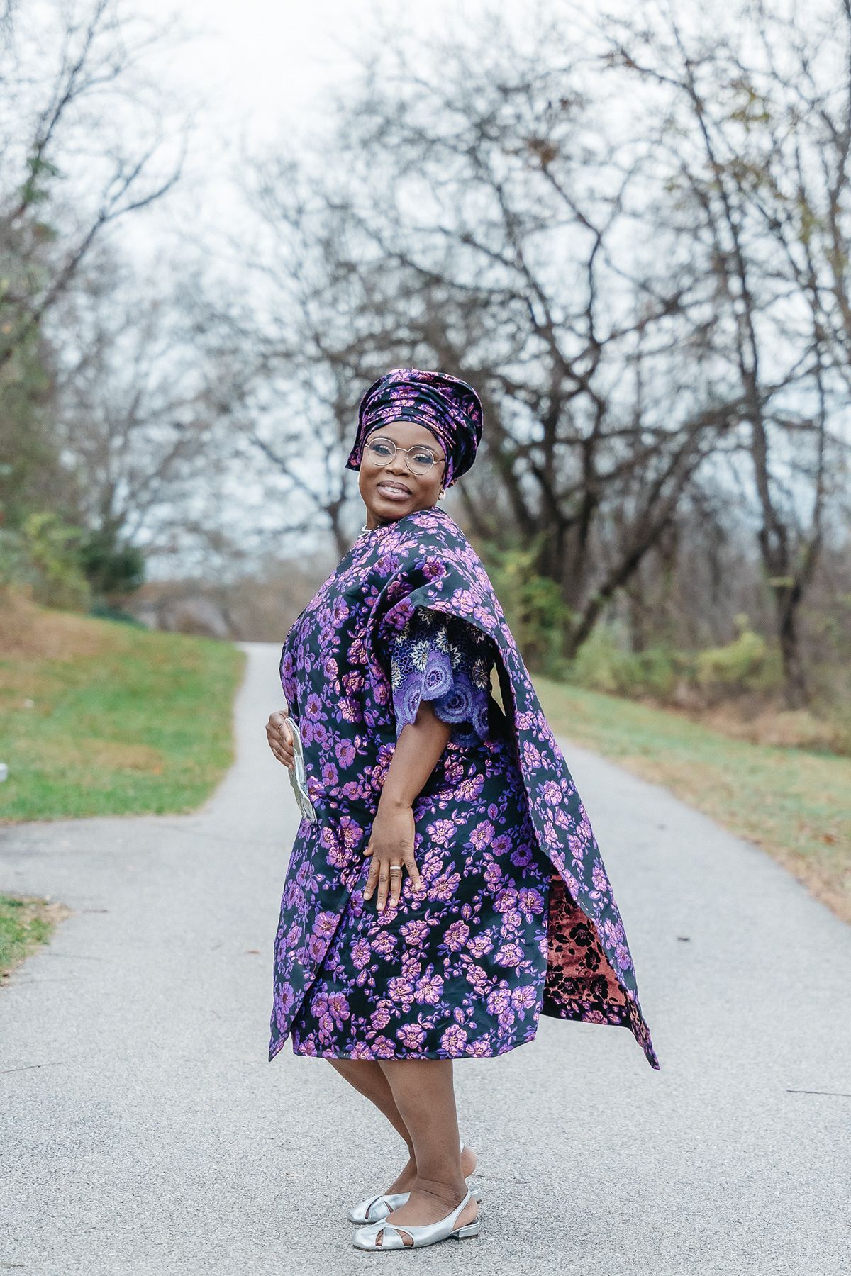 A person twirling in a purple floral dress and matching headwrap on a park path.