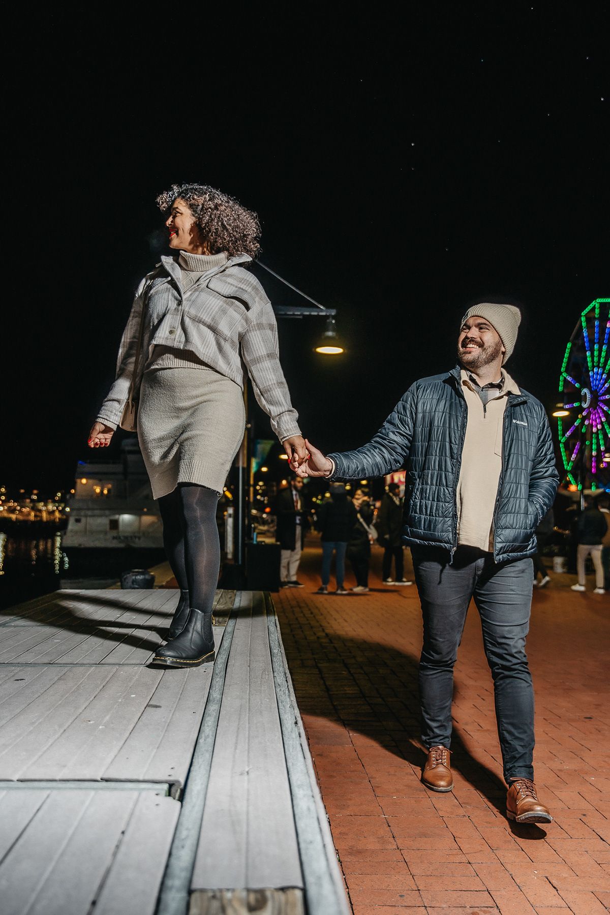A couple holds hands while walking near a carnival at night with a Ferris wheel in the background.