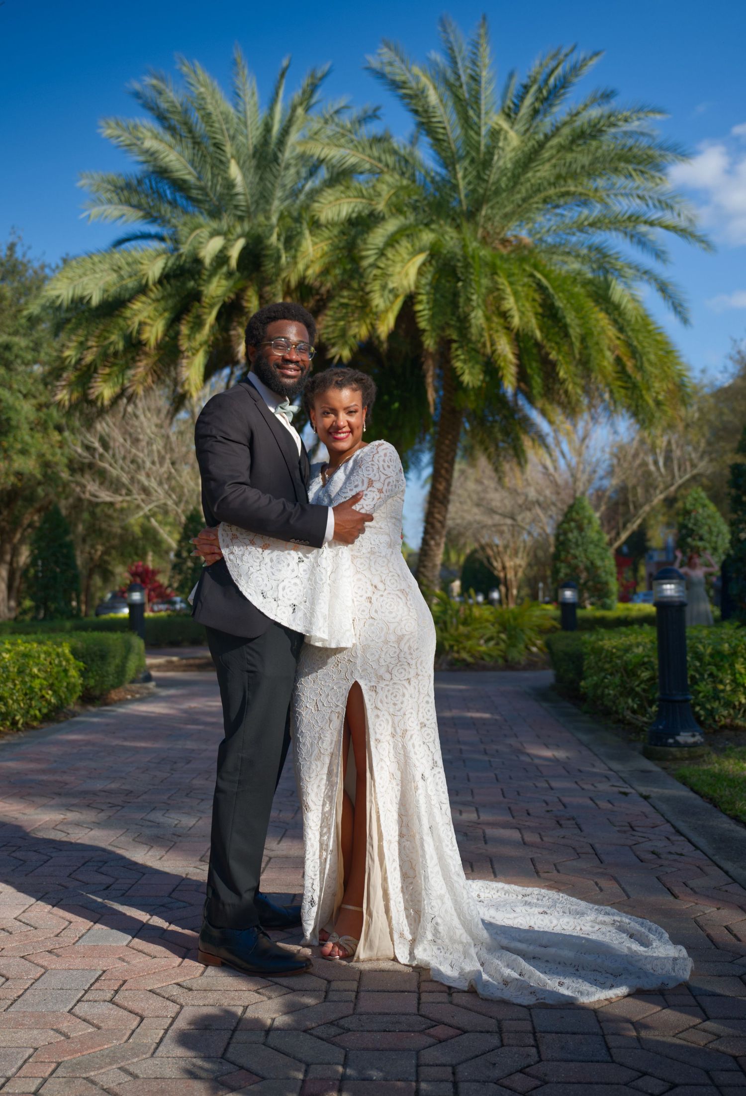 Couple embracing under palm trees on brick pathway during wedding photoshoot.