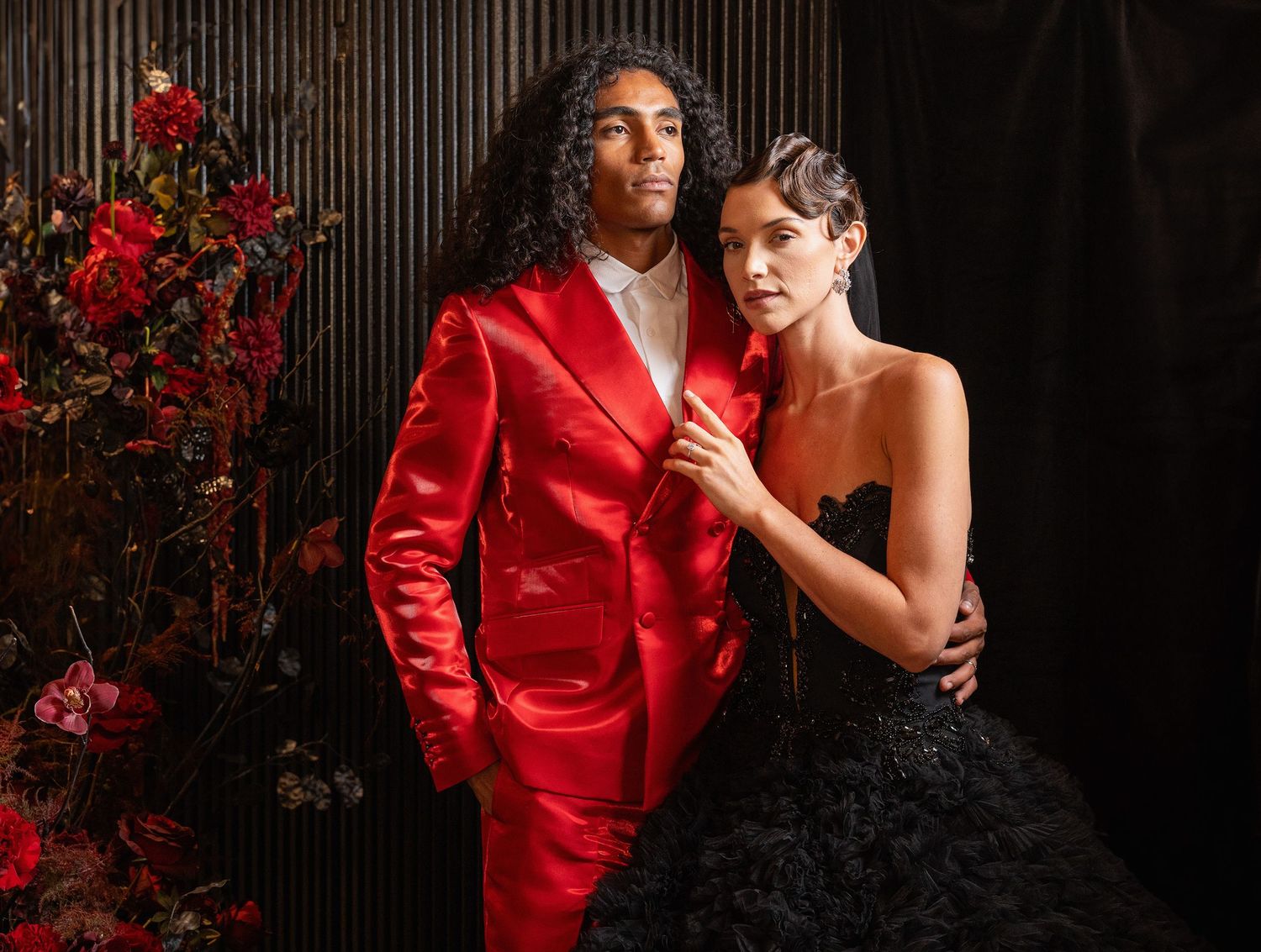 A couple in dramatic formal wear posing against dark backdrop with red floral arrangements.