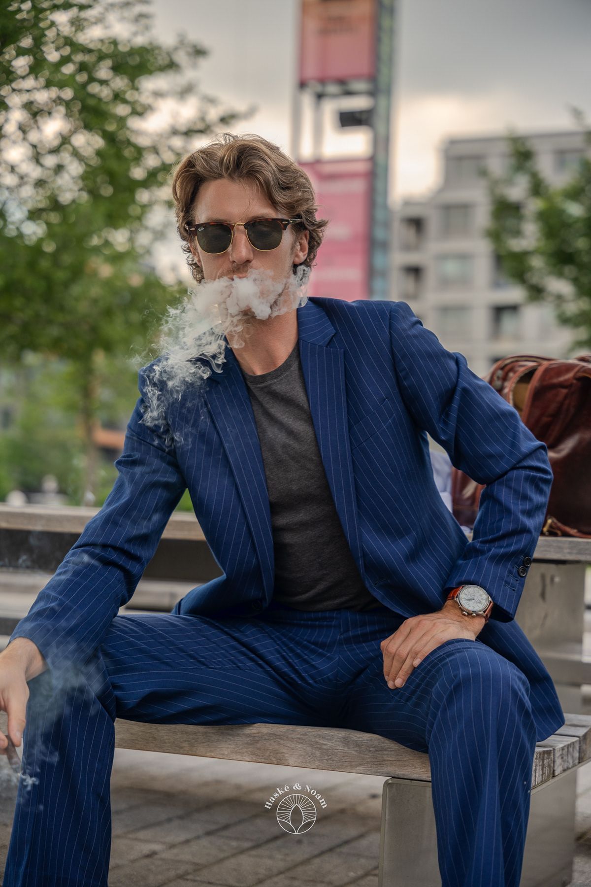 Stylish person in pinstripe suit exhaling smoke while sitting on bench in urban setting.