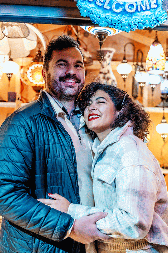 Couple embracing under festive blue welcome sign with warm lighting.