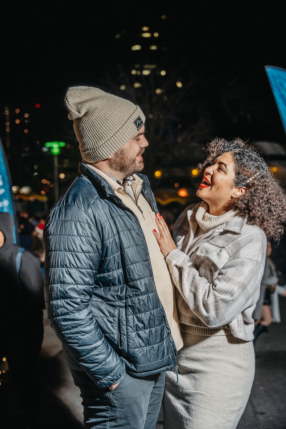 Nighttime portrait of a couple laughing together at an outdoor event with city lights behind them.