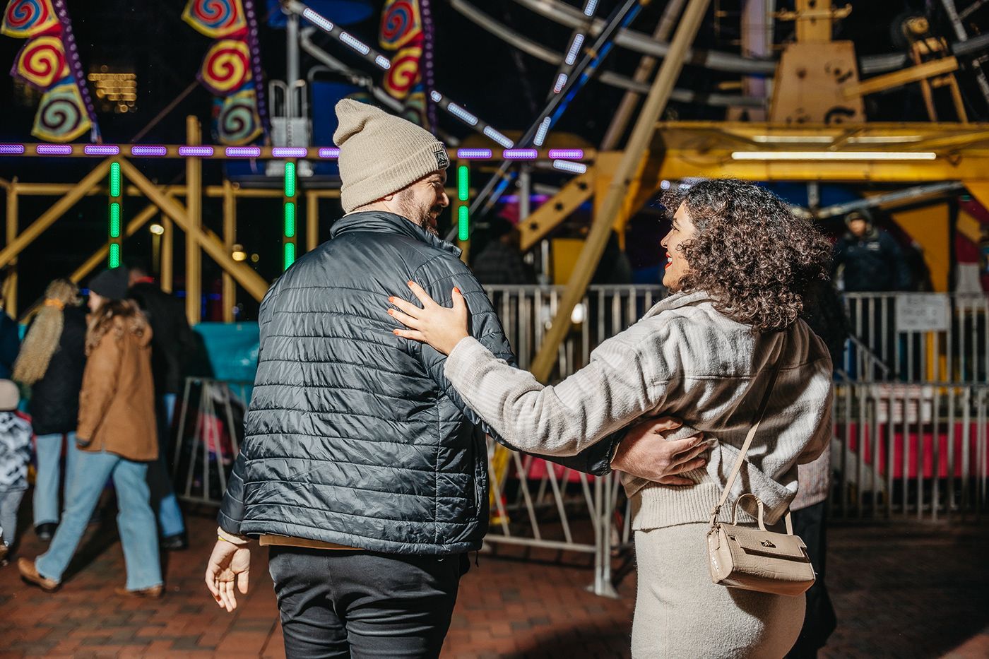 A couple walks together at a carnival with festive lights and rides in the background.