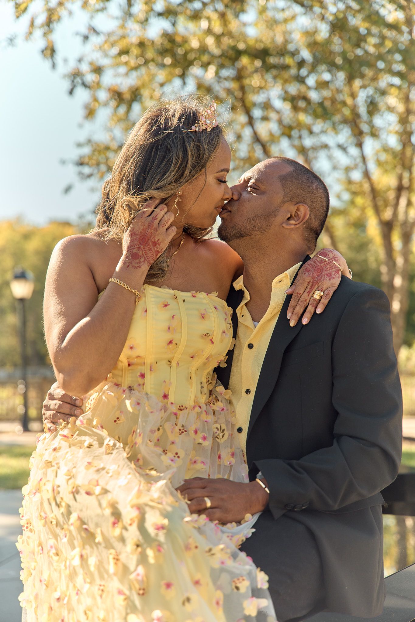 A romantic kiss shared between a couple in formal wear during a sunny outdoor photoshoot.