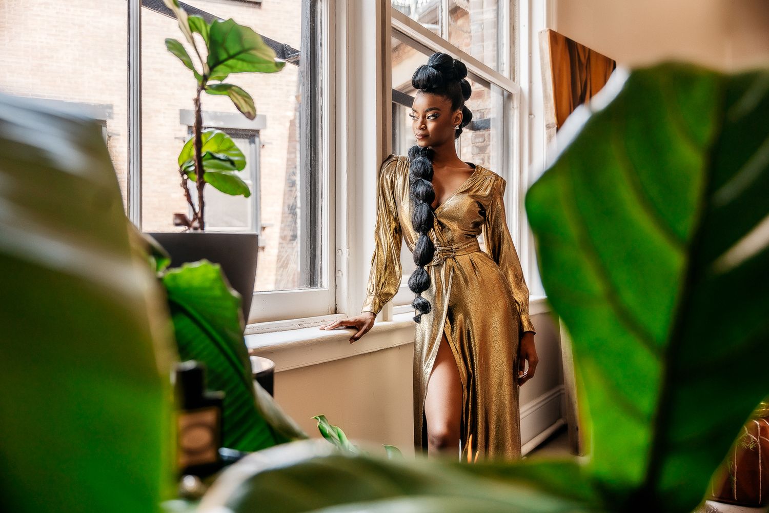Fashion photo of gold dress between tropical plants by sunlit window.