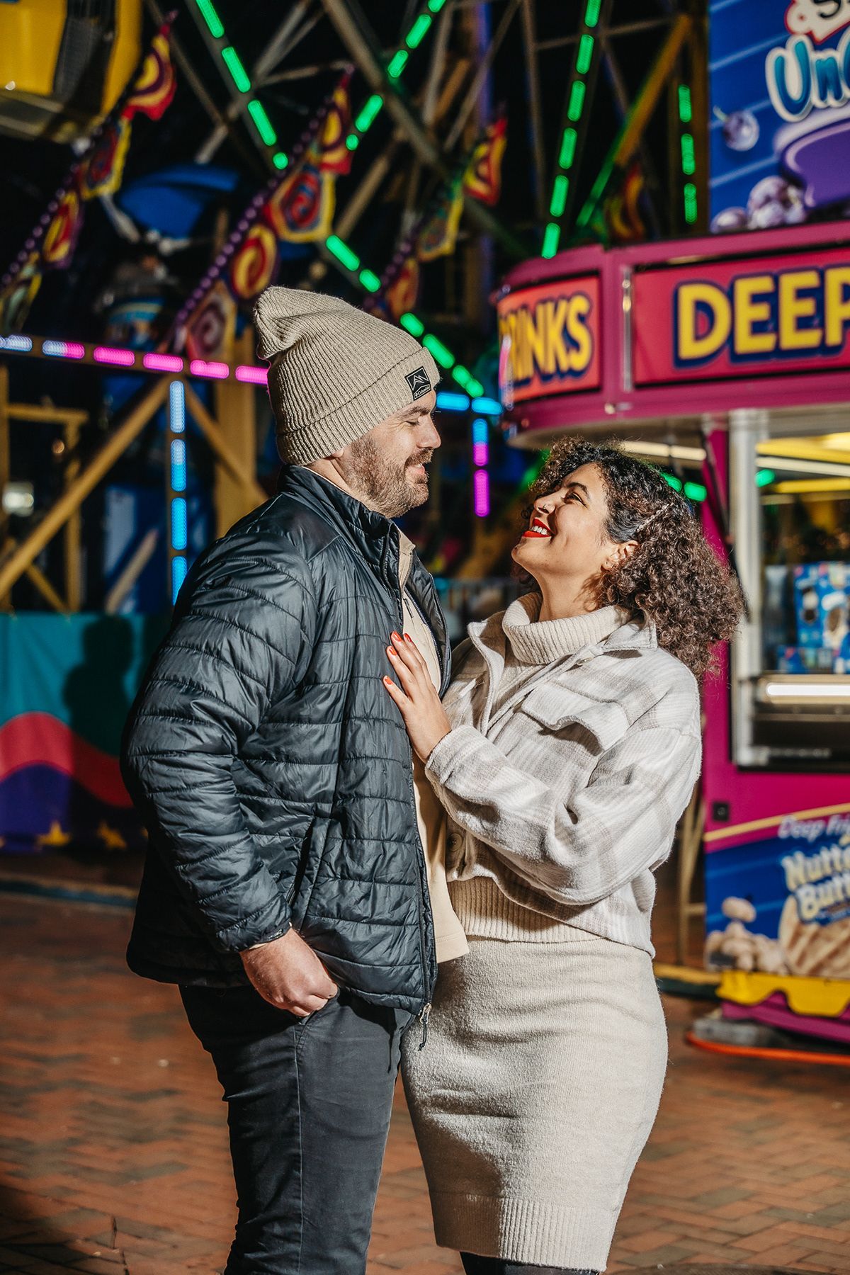 A couple shares a tender moment at a carnival with colorful neon lights in the background.