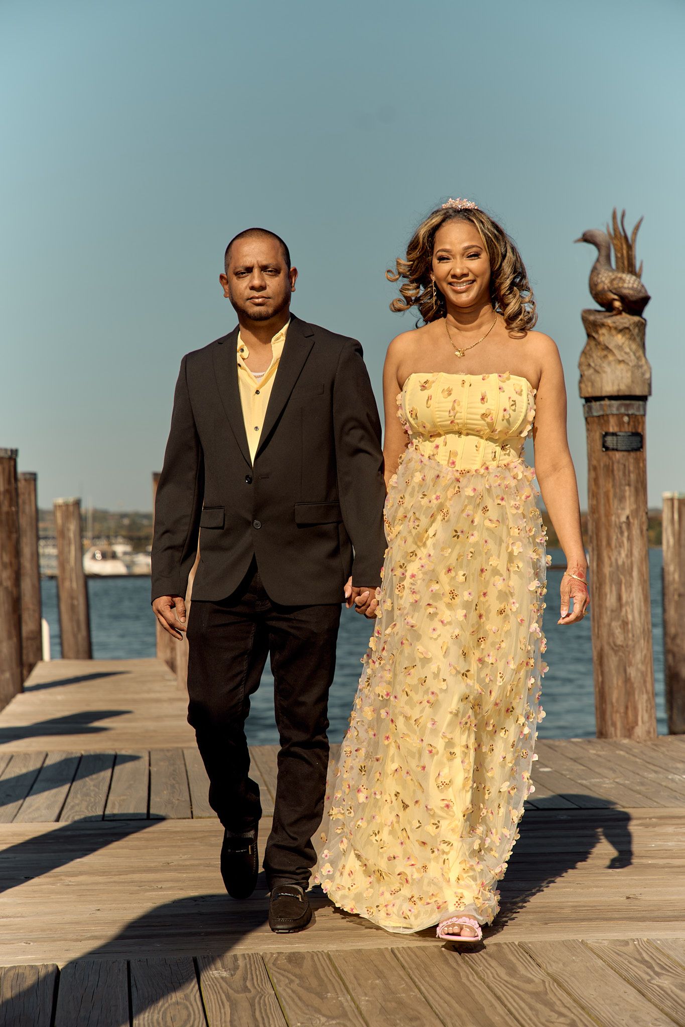 A couple walks hand in hand on a wooden dock, with the woman wearing a yellow floral gown and the man in a dark suit.