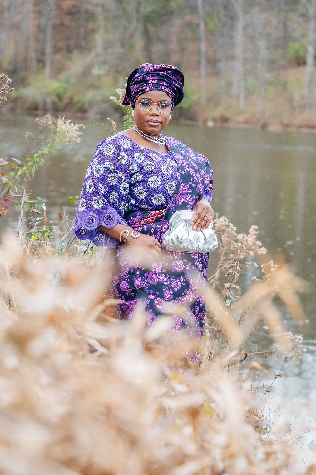 A portrait by a lake featuring someone in a purple patterned dress and headwrap holding a silver clutch.